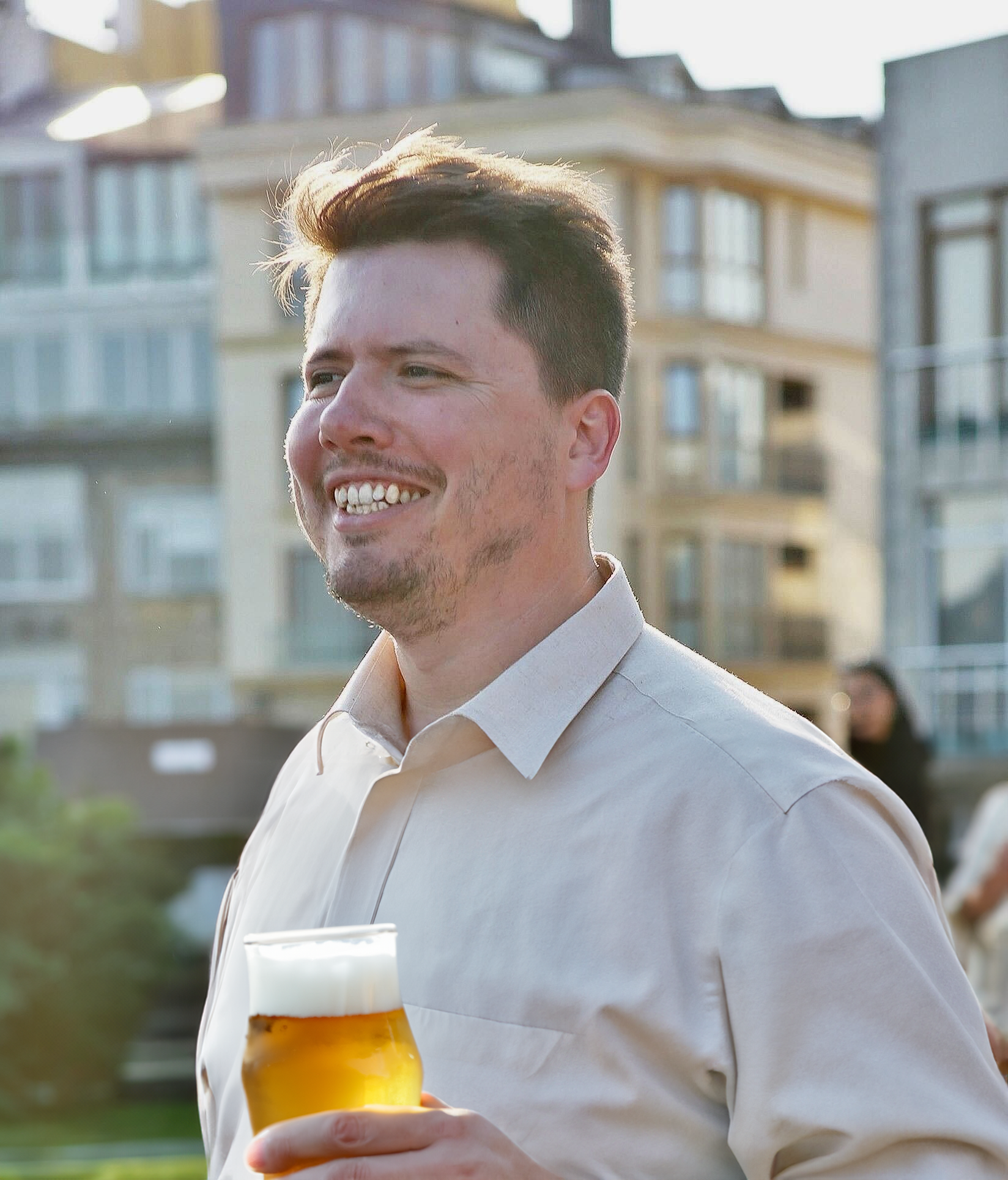 A man smiling and holding a glass of beer outdoors in front of modern apartment buildings.