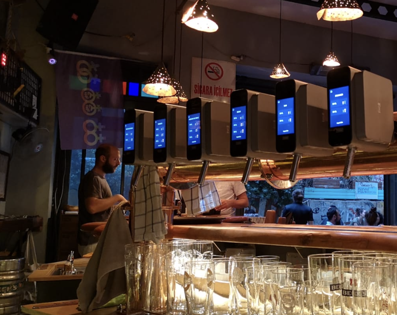 Bar with multiple electronic ordering screens hanging from the ceiling, a bartender preparing drinks, and patrons visible through the open window in the background.