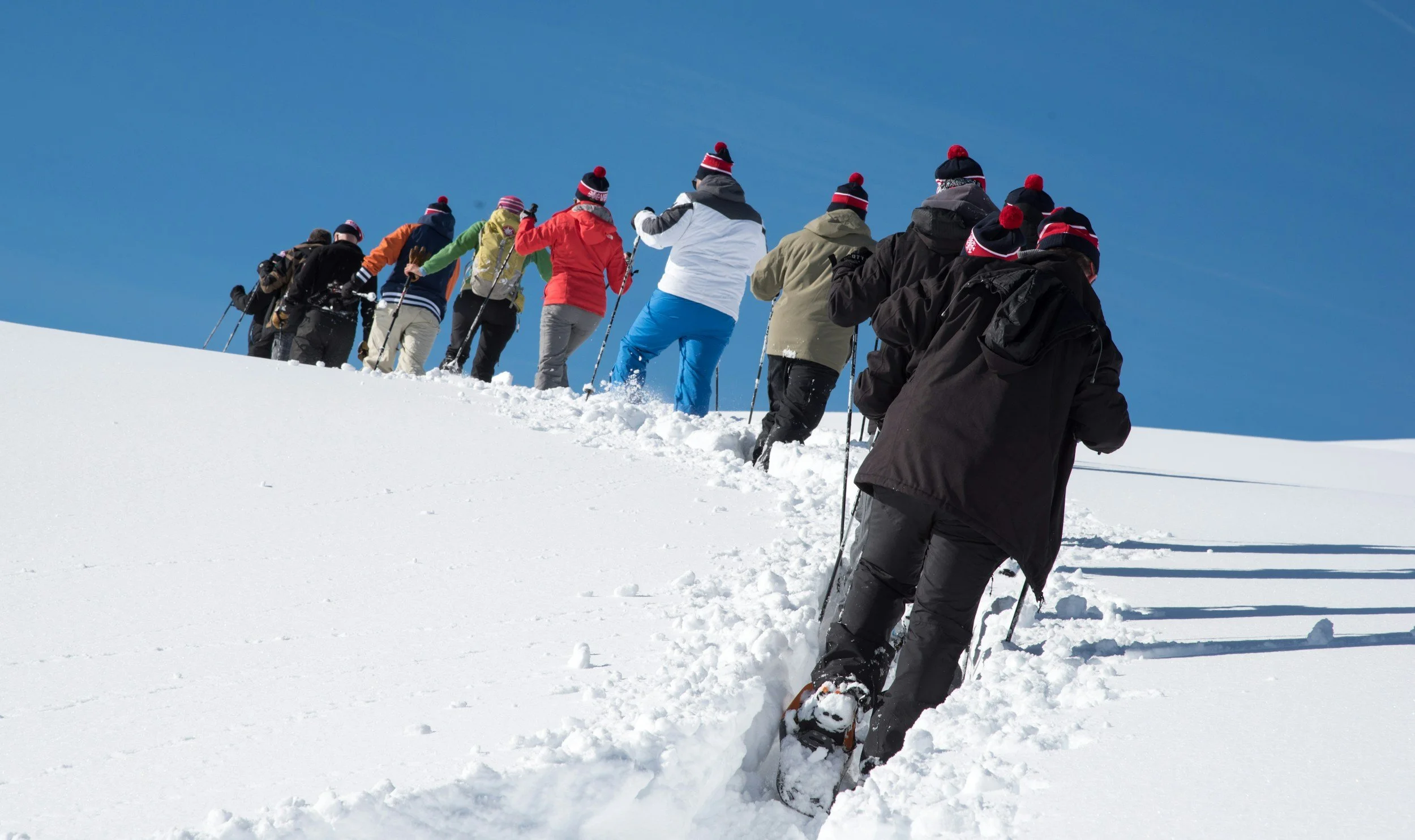 A group of people wearing winter clothing and hats with pom-poms hiking up a snowy slope during daytime under clear blue skies.
