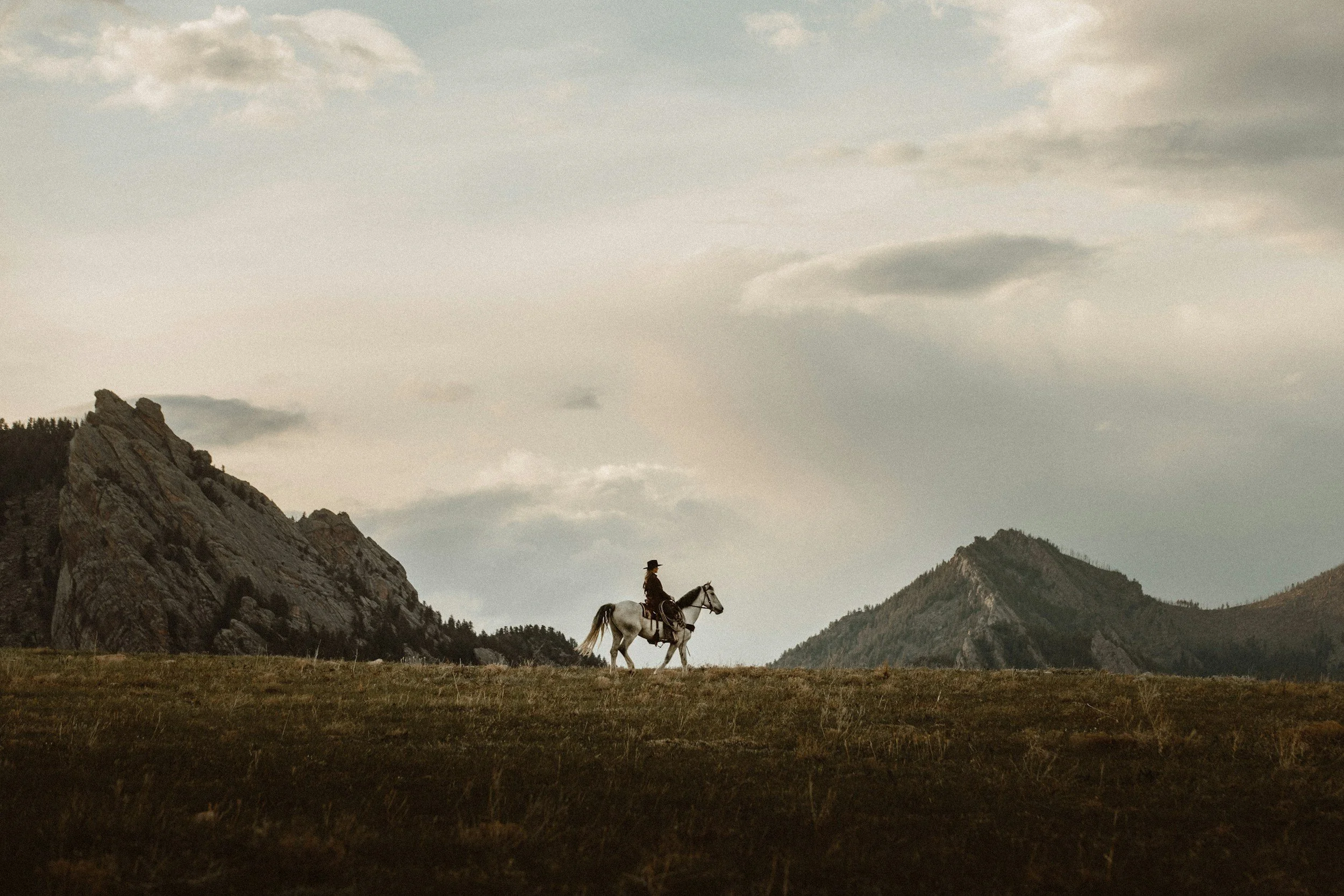 A person riding a white horse across a vast open field with mountains in the background and a cloudy sky overhead.
