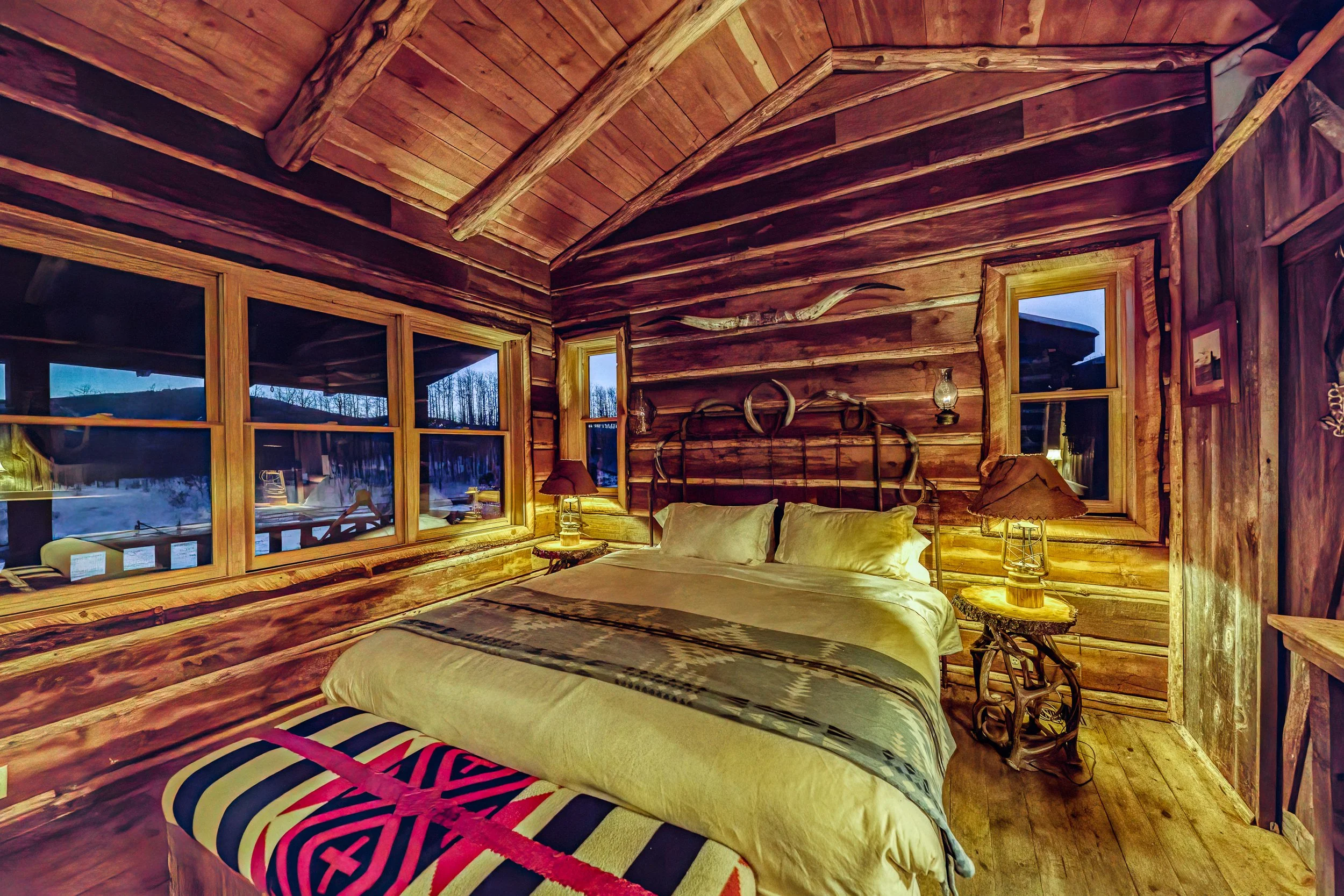 A rustic bedroom with wooden walls, ceiling, and floors, illuminated by warm lighting from lamps on nightstands on each side of the bed. The bed has white bedding and pillows, and there is a colorful striped bench at the foot of the bed. Large window