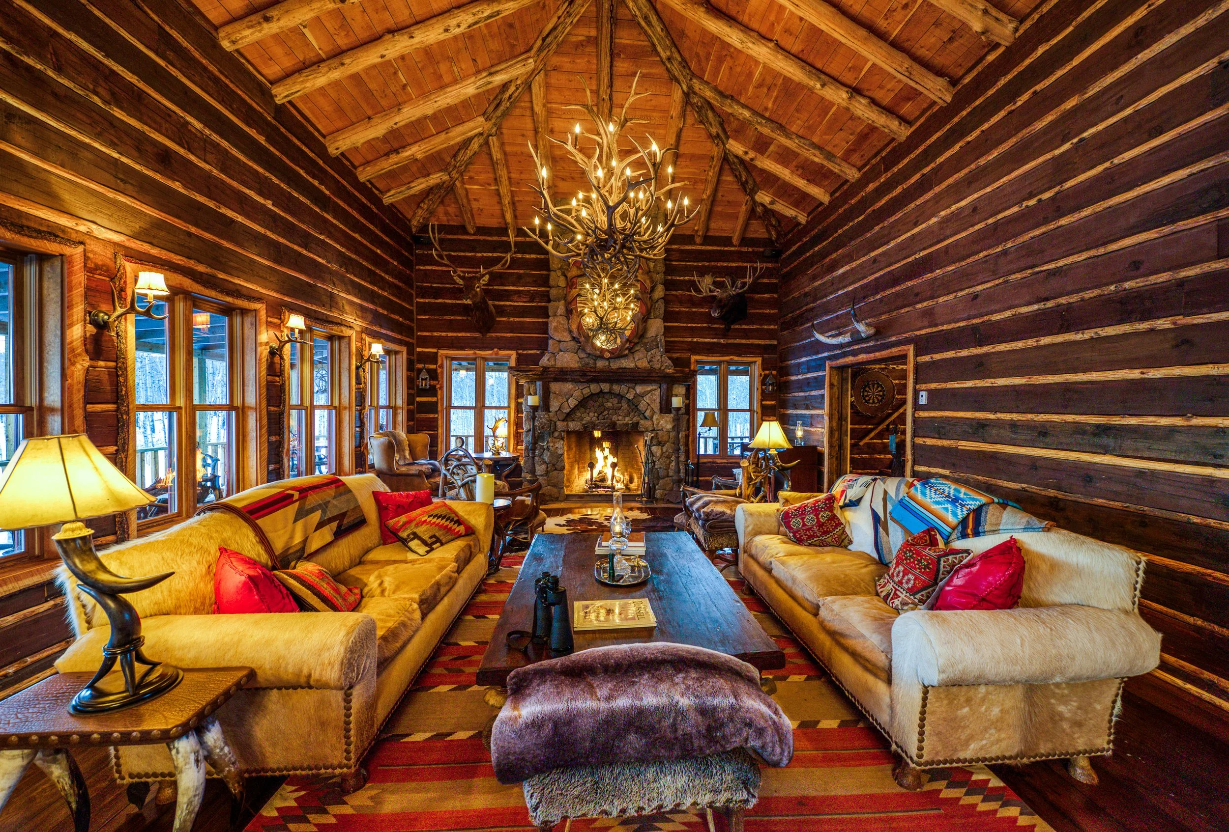 Interior of a rustic log cabin living room with wooden walls, a stone fireplace with a fire, and a chandelier made of antlers and branches. There are two sofas with patterned and red cushions, a large wooden coffee table, and various lamps and chairs
