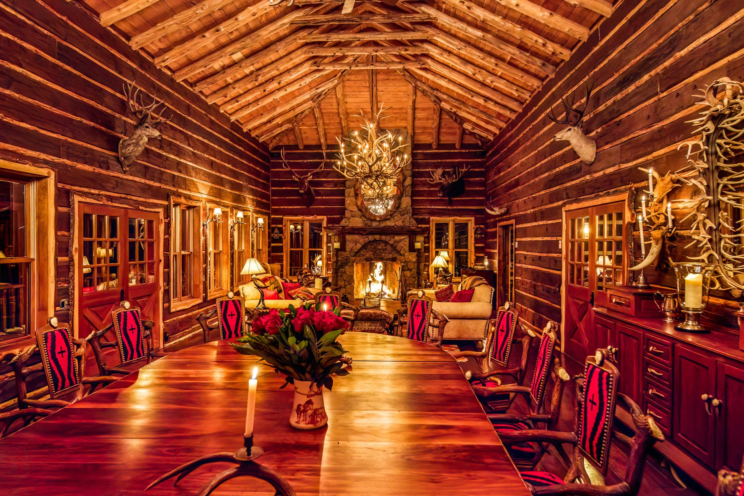 A rustic dining room with a long wooden table, surrounded by wooden chairs with red and black striped upholstery. The room has log cabin walls decorated with mounted deer heads and a large chandelier made of antlers hanging from a peaked wooden ceili
