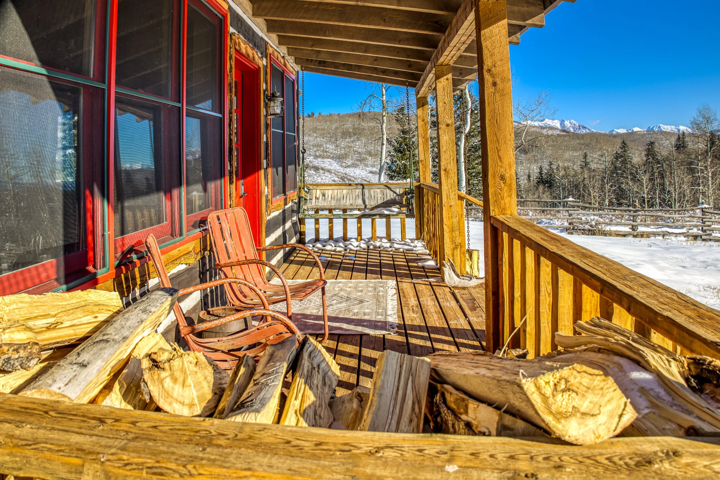 Wooden porch of a cabin with snow, red window frames, chairs, and a swing, with mountains and trees in the background on a clear winter day.