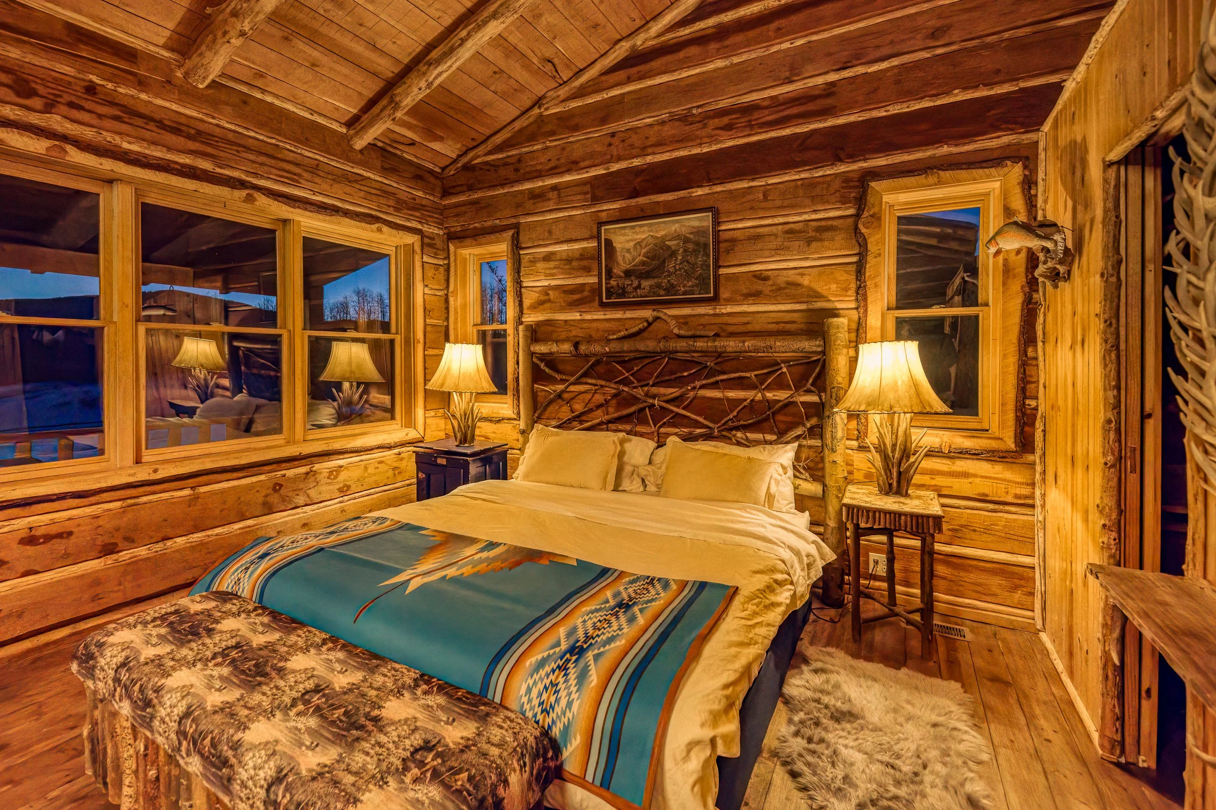 Cozy bedroom in a log cabin with wooden walls, a bed with beige bedding and a decorative blanket, three lamps on side tables, and framed artwork above the bed.