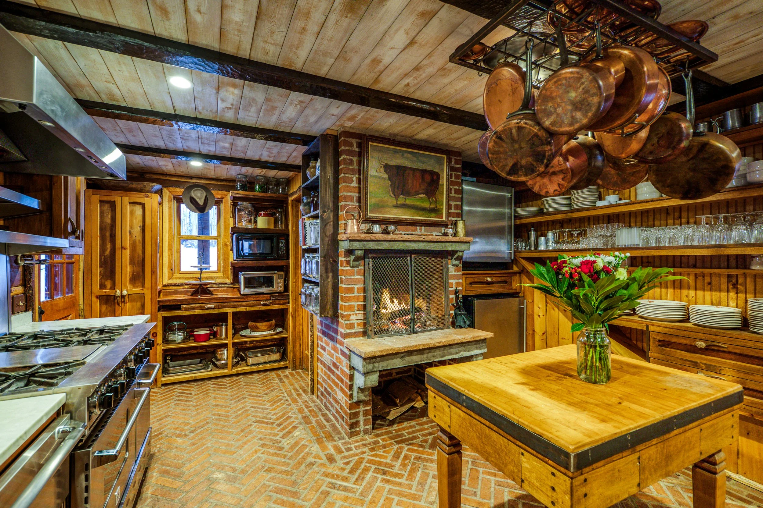Interior view of a cozy rustic kitchen with brick and wooden walls, hanging copper pots, a fireplace, a wooden table with a vase of red flowers, and various kitchen appliances and utensils.