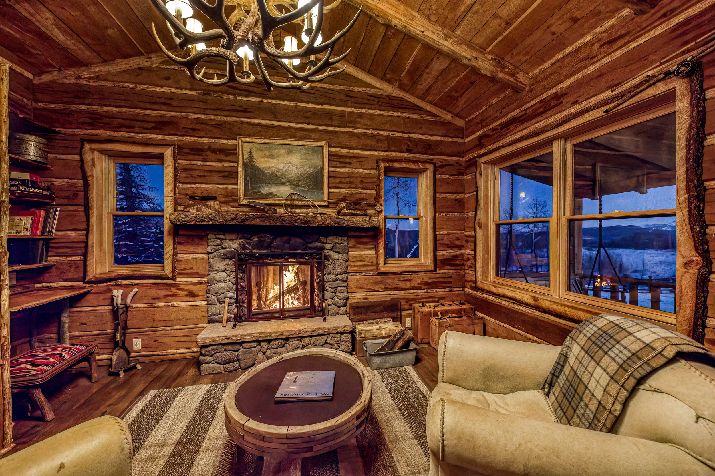 Cozy living room with wood-paneled walls, a stone fireplace with a lit fire, and large windows showing a winter landscape outside. There is a cushioned armchair, a round coffee table, and a ceiling chandelier with antler design.