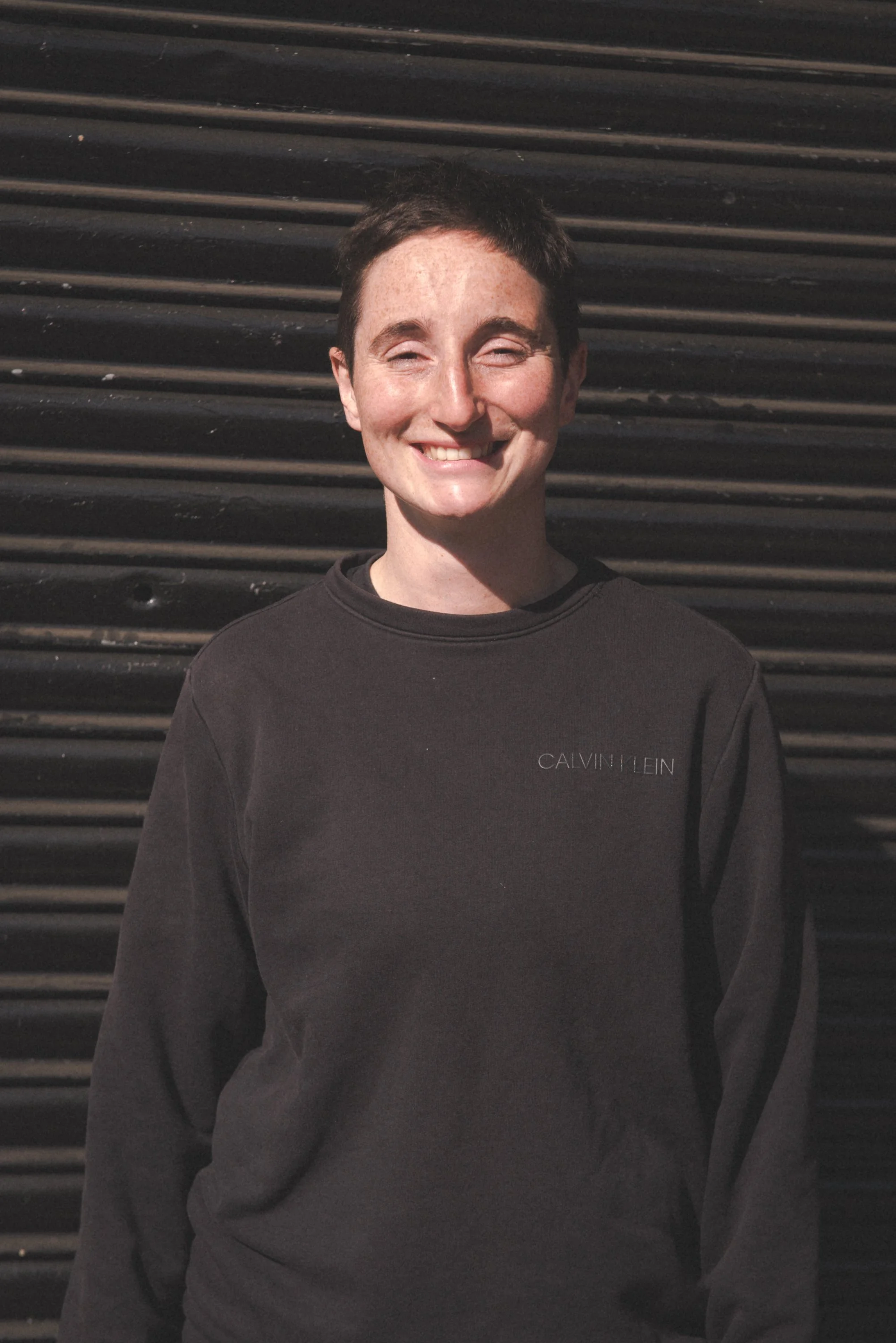 A person with short dark hair smiling with closed eyes, wearing a black Calvin Klein sweatshirt, standing in front of a black corrugated metal wall.