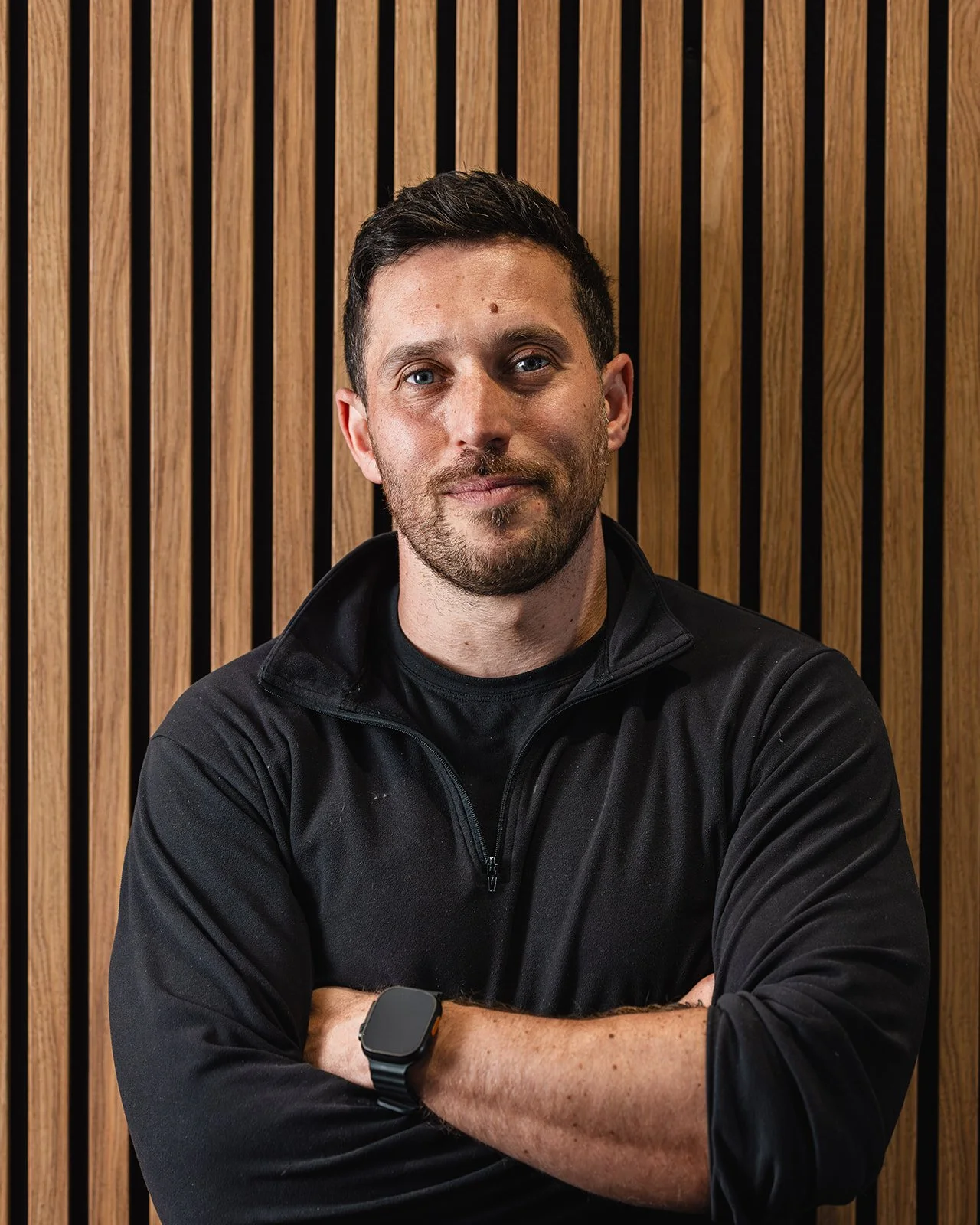 A man with dark hair and a beard, wearing a black zip-up jacket and smartwatch, standing with arms crossed in front of a wooden slat wall.