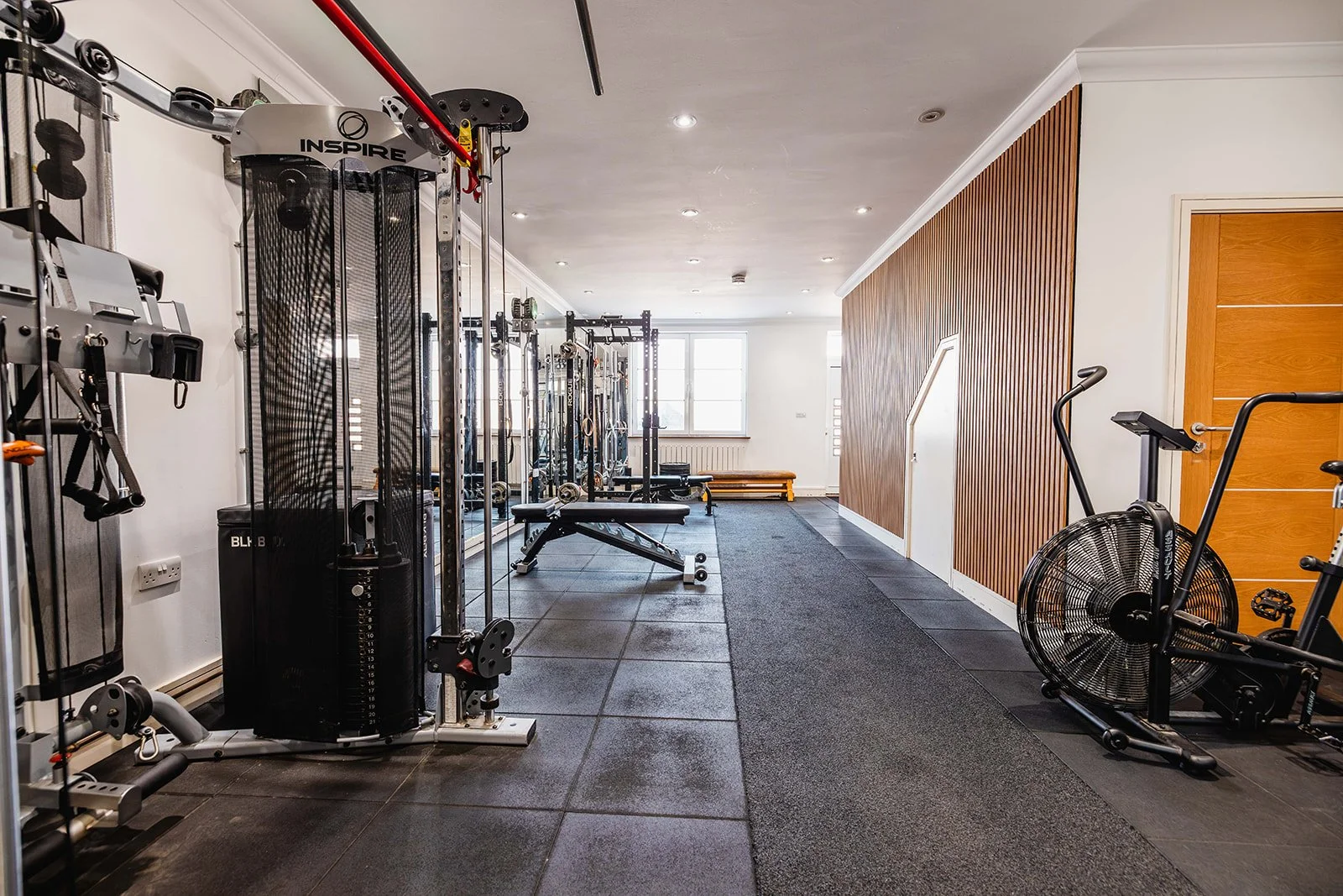 A well-equipped home gym with various exercise machines, including a large multi-purpose fitness station, a bench, and an exercise bike with a fan wheel, all set against white walls, a wooden accent wall, and windows at the far end.