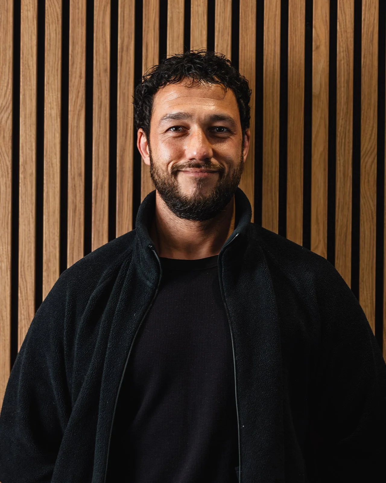 Portrait of a man with curly dark hair and a beard, smiling, wearing a black jacket and black shirt, standing against a wooden slat background.