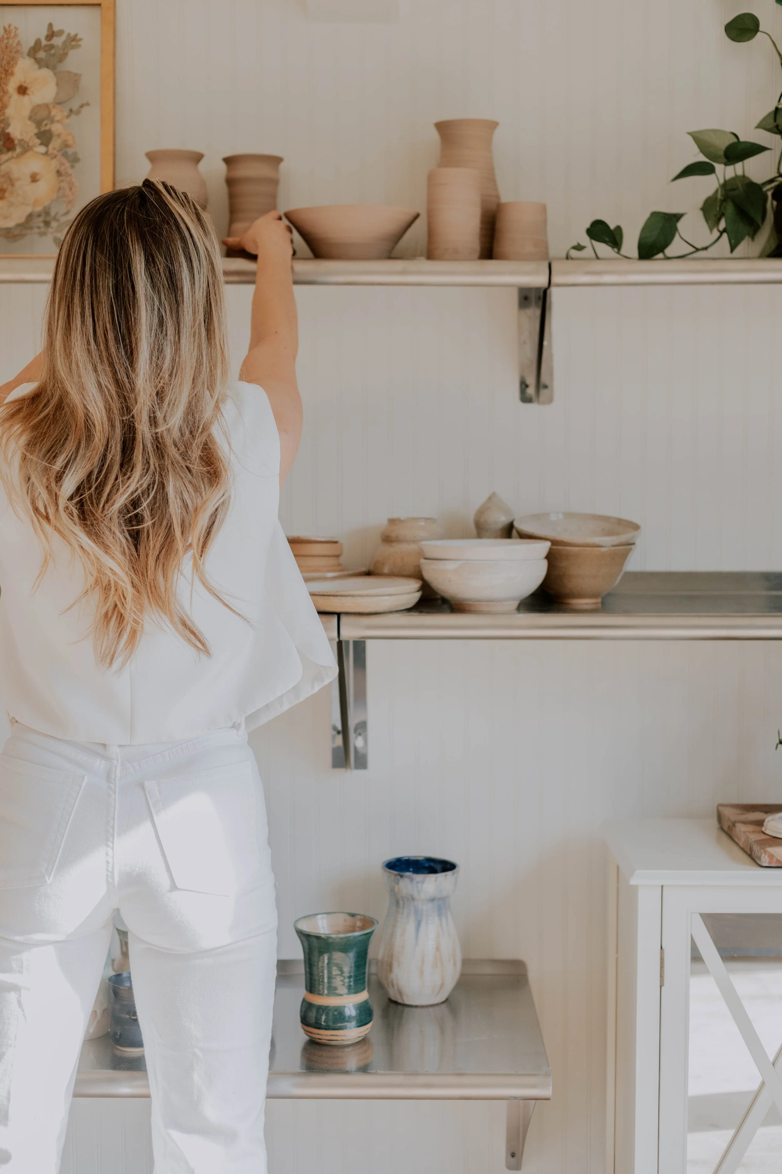 Person with long, wavy blonde hair wearing white clothing, pointing at pottery on shelves in a pottery studio with bowls, vases, and plants.