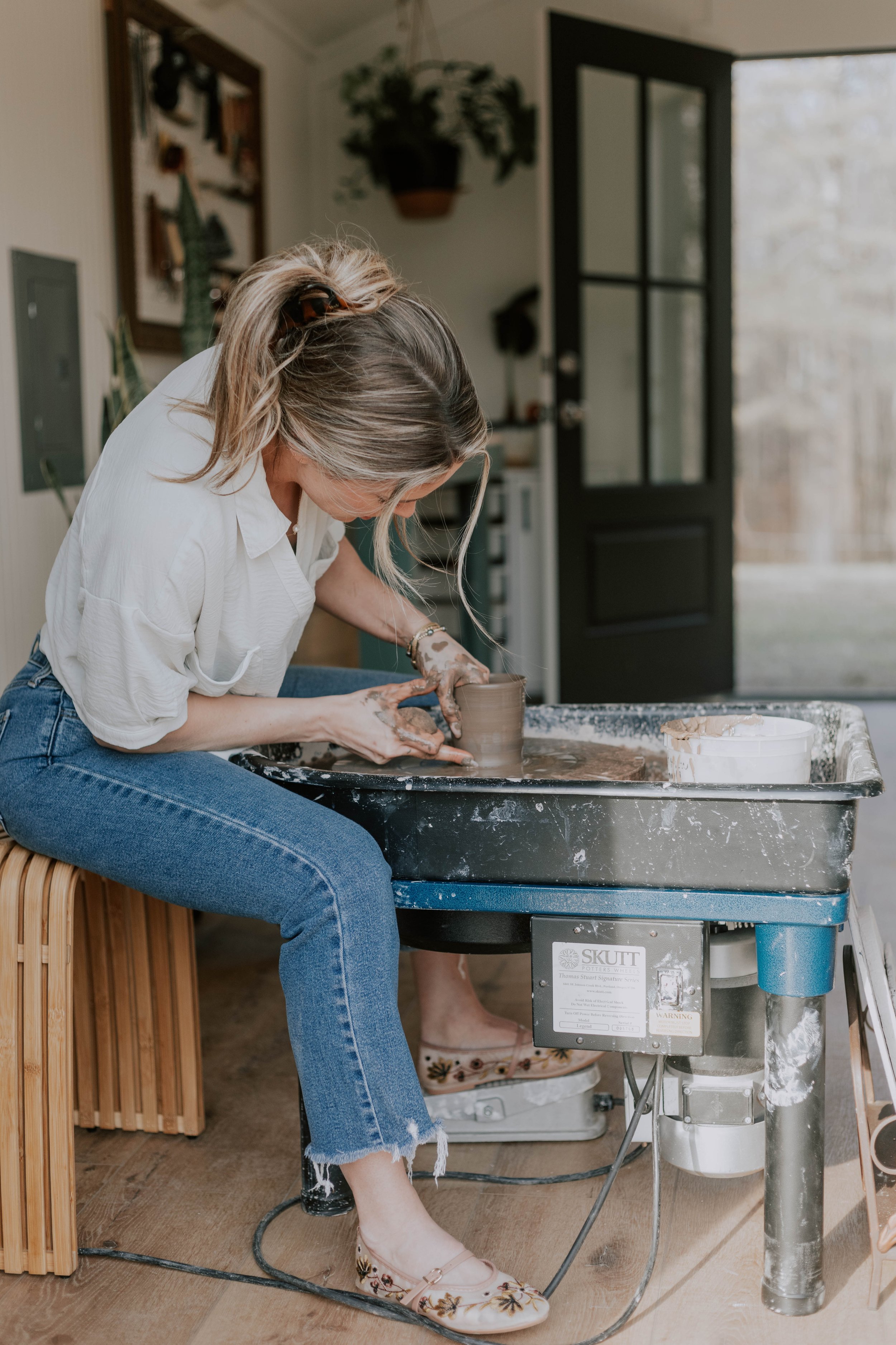 A woman with blonde hair tied in a ponytail, wearing a white shirt and blue jeans, is pottery making on a pottery wheel in a cozy indoor space with a wooden floor and plants in the background.