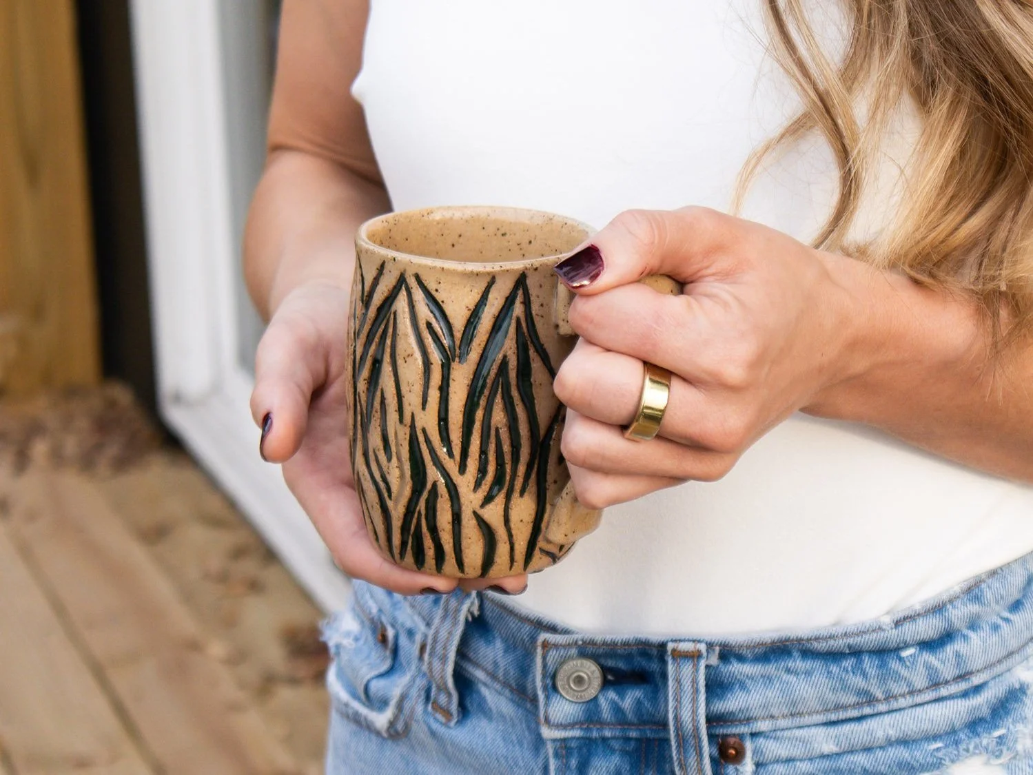Person holding a ceramic mug with a leaf pattern, wearing a gold wedding band and a beige short-sleeve shirt, standing indoors near a door.