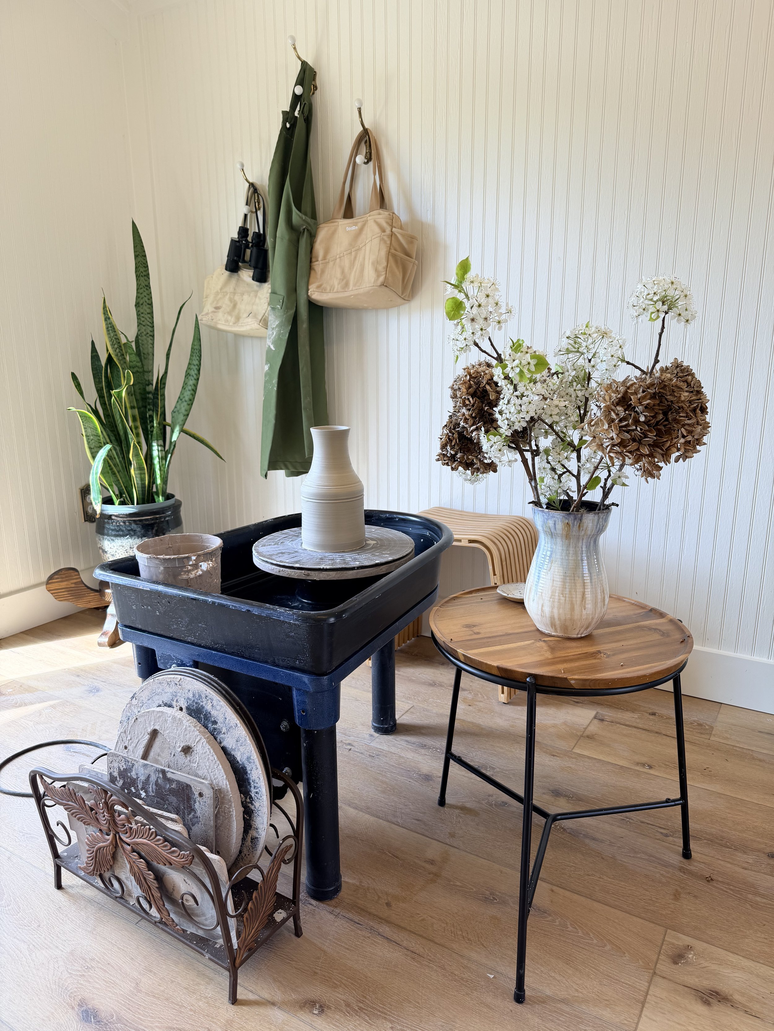 A room with wooden flooring and white paneled walls, featuring potted plants, a small wooden side table with a vase of flowers, a black cart with pottery, and hooks on the wall holding bags and umbrellas.