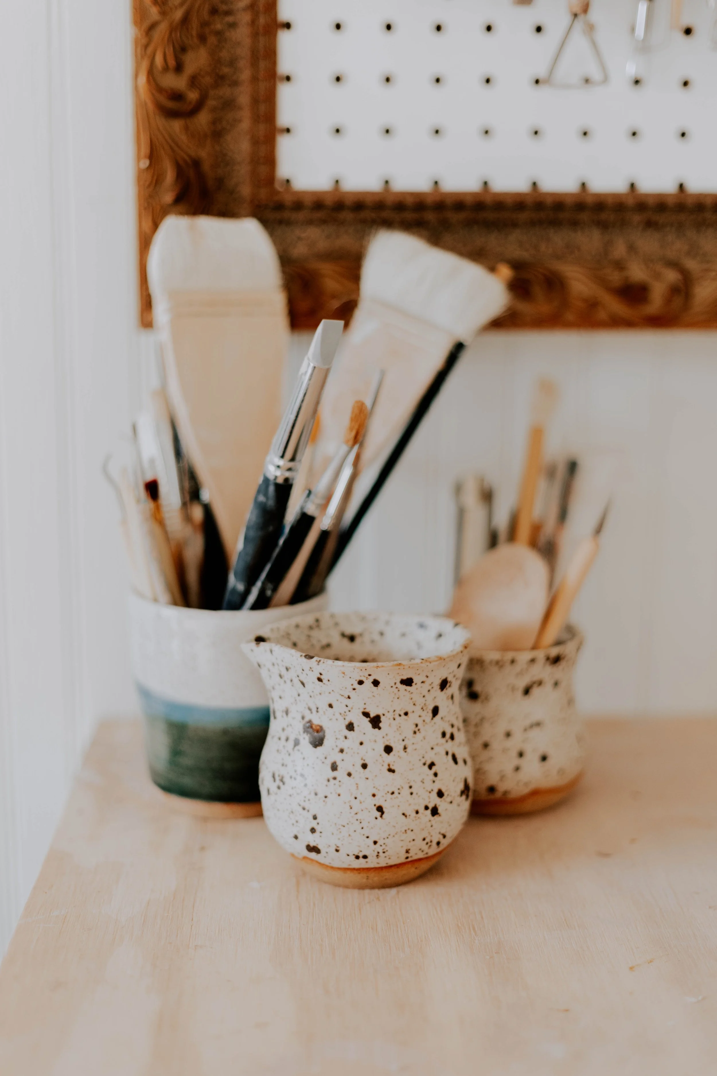 Three handmade ceramic vases with paintbrushes inside, placed on a wooden surface, with a pegboard and wooden frame in the background.