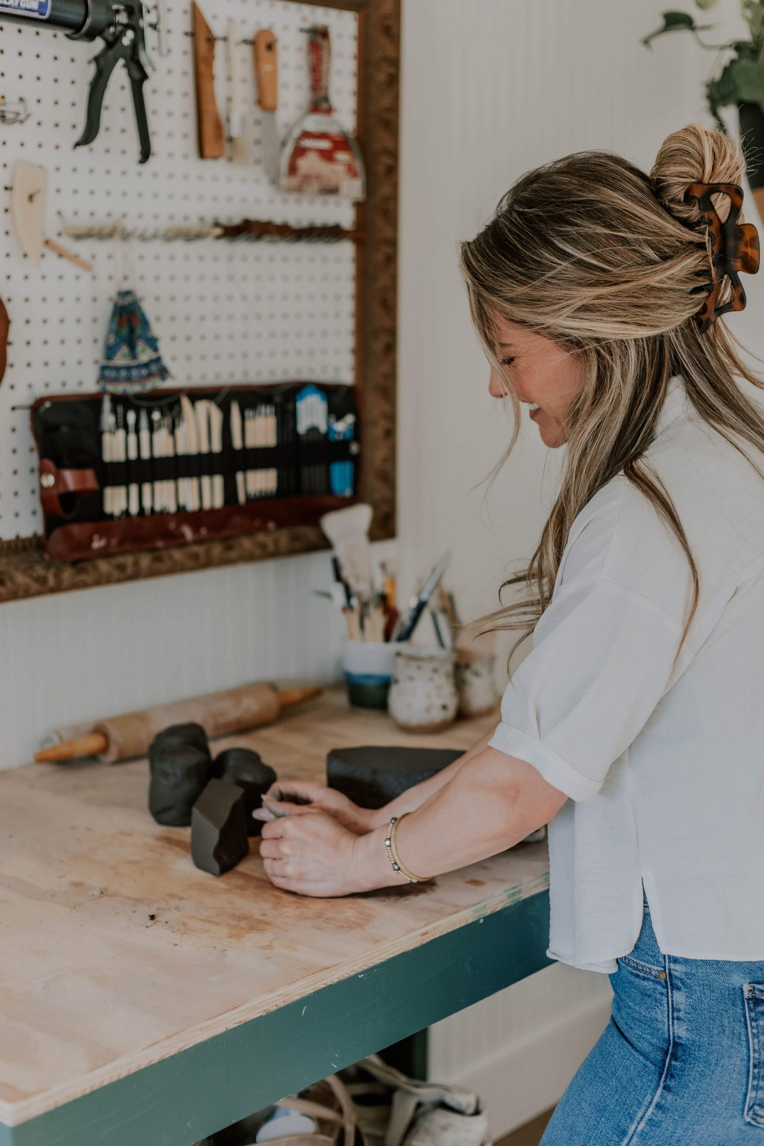 A woman with blonde hair in a bun, wearing a white shirt and blue jeans, working at a wooden workbench with sculpting tools and black clay, in a workshop with a pegboard holding tools in the background.