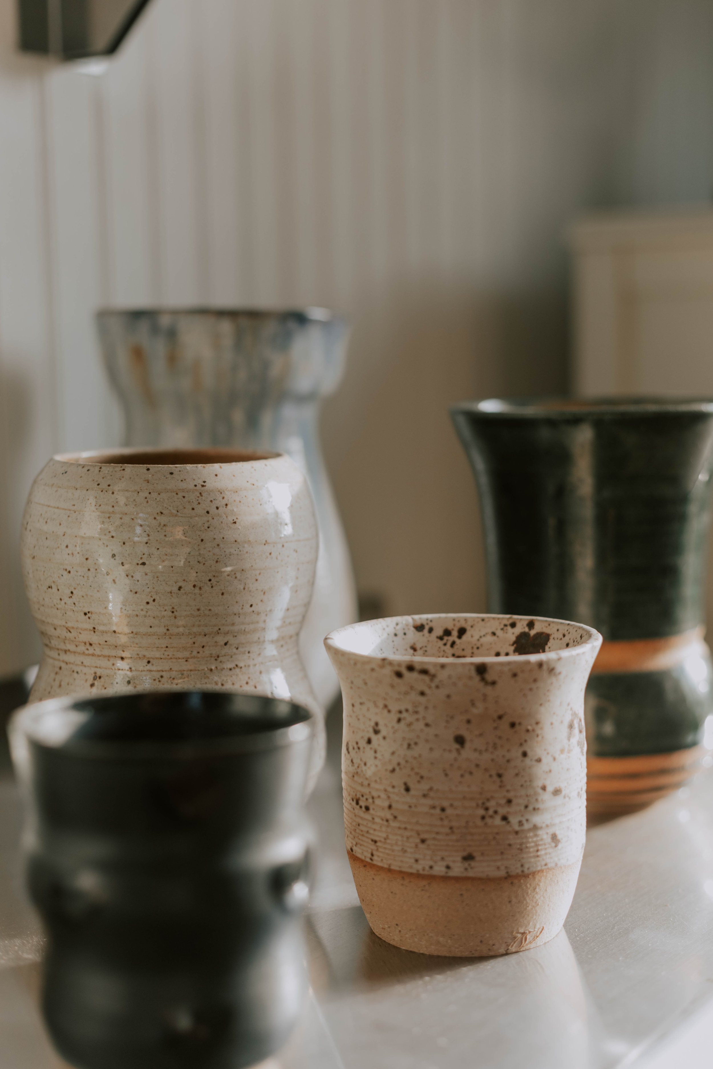Close-up of ceramic vases and cups on a surface, with a cream-colored vase featuring dark specks and a larger vase in the background, all with a blurred neutral background.