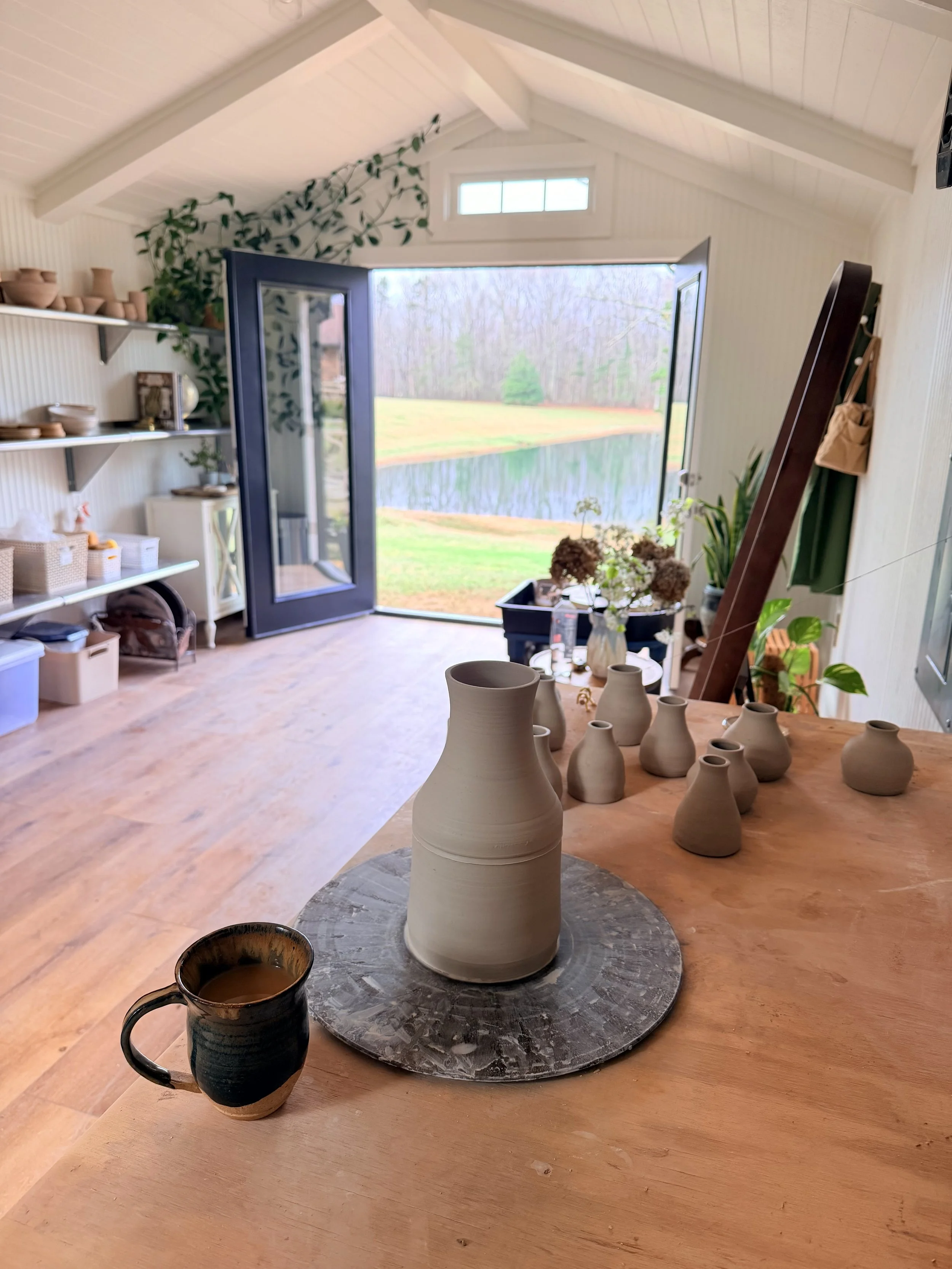 Interior view of a room with open window overlooking a lake and forest, cozy and decorated with pottery and plants.