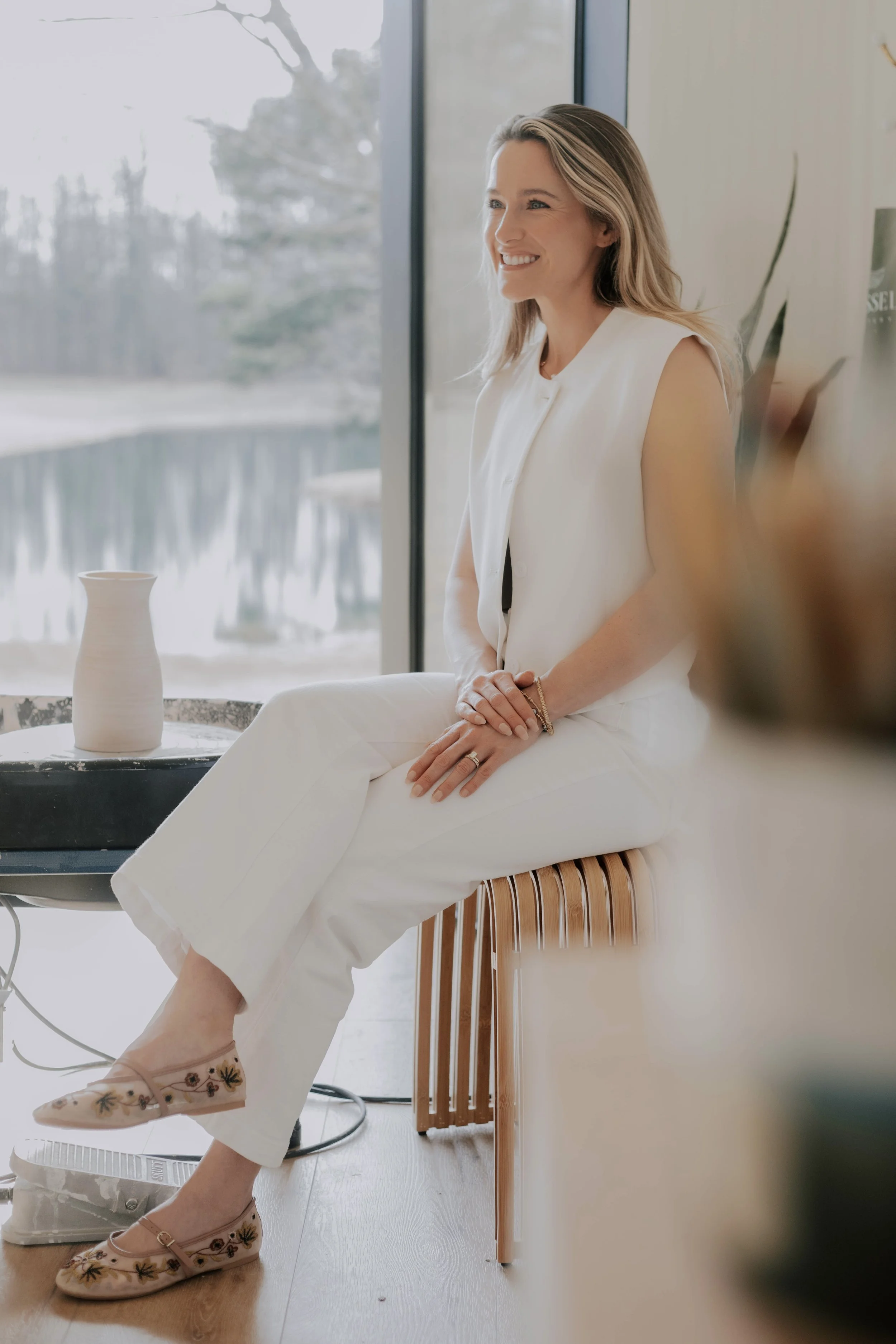 A woman with blonde hair smiling, sitting on a wooden chair near a large window with a snowy outdoor landscape in the background. She is wearing white pants, a sleeveless white top, and floral embroidered shoes. A ceramic vase is on a table nearby.