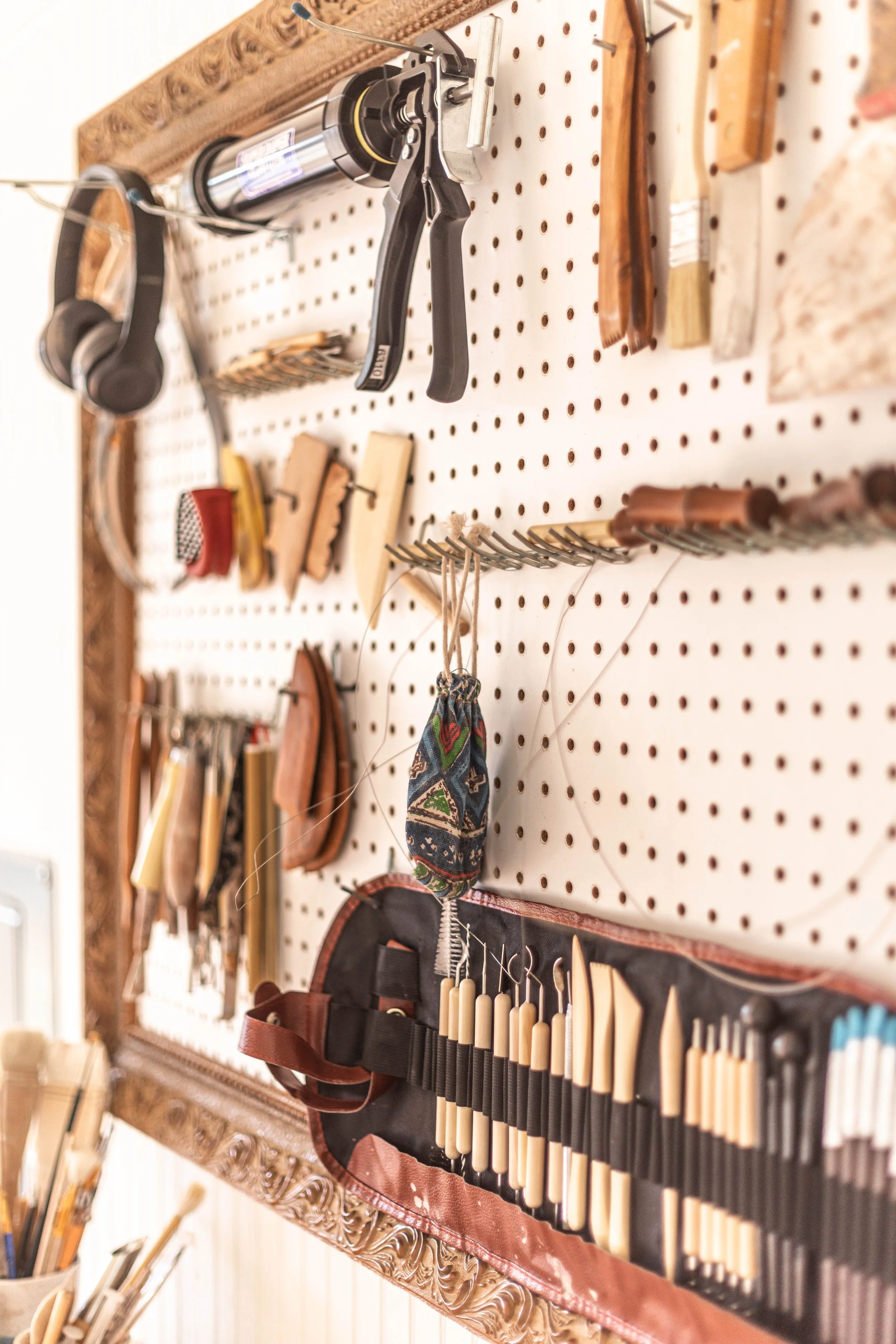 A wall-mounted pegboard with various tools and accessories: a vise grip clamp, a small tin, wooden handled brushes, and a decorated cloth pouch holding scissors.