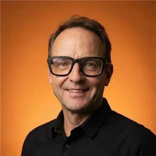 Porträt eines lächelnden Mannes mit schwarzen Brillen vor orangem Hintergrund.