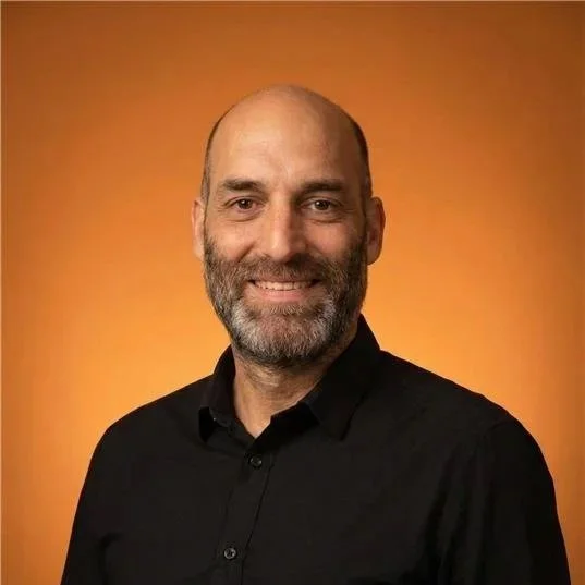 Porträt eines lächelnden Mannes mit Bart und schwarzem Hemd vor orangem Hintergrund.