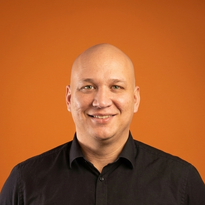 Porträt eines Mannes vor einem orangefarbenen Hintergrund, lächelnd, mit schwarzem Hemd.