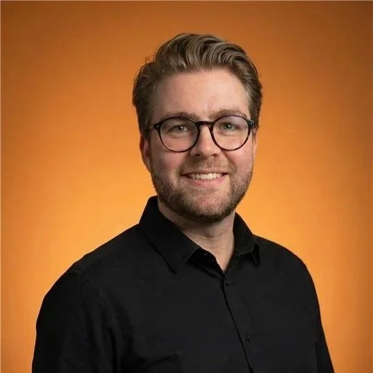 Ein Mann mit Brille, dunklem Hemd, lächelnd, vor orangem Hintergrund.