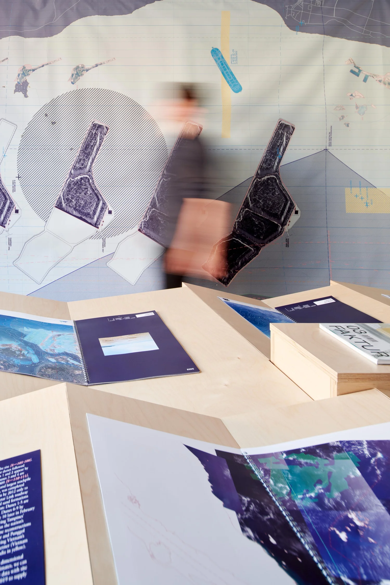 Exhibition view of Territories of Territory Extraction featuring a wall graphic of island forms, a visitor walking by, and books displayed on stepped wood platforms.