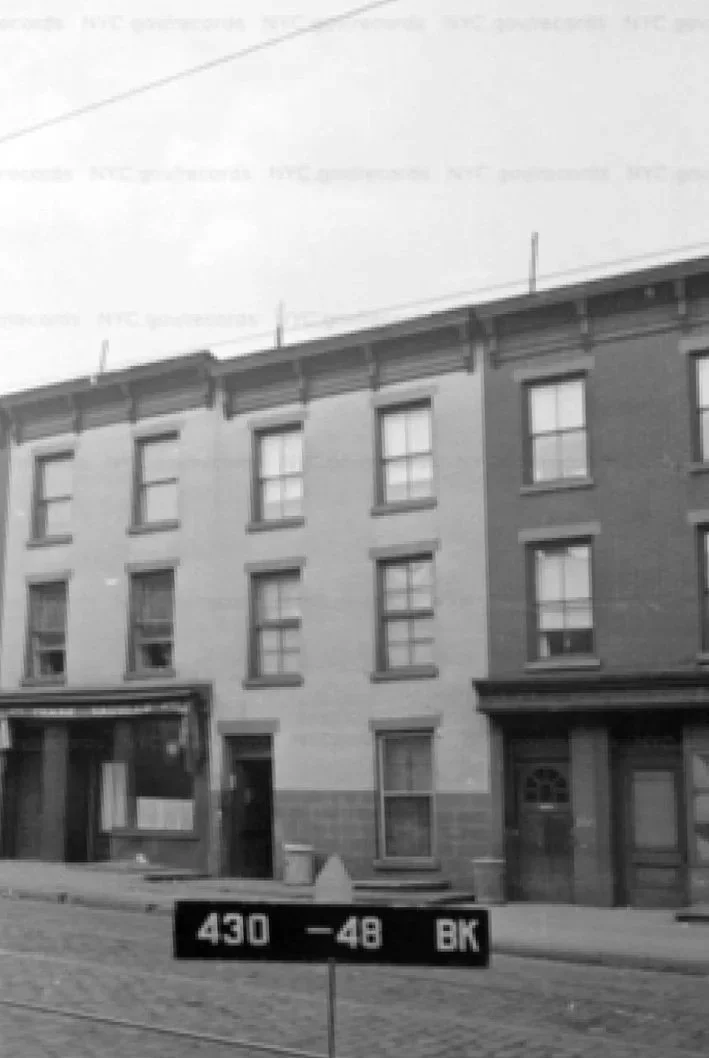 Black-and-white photo of three attached brick townhouses with flat roofs and simple façades, taken in Brooklyn.