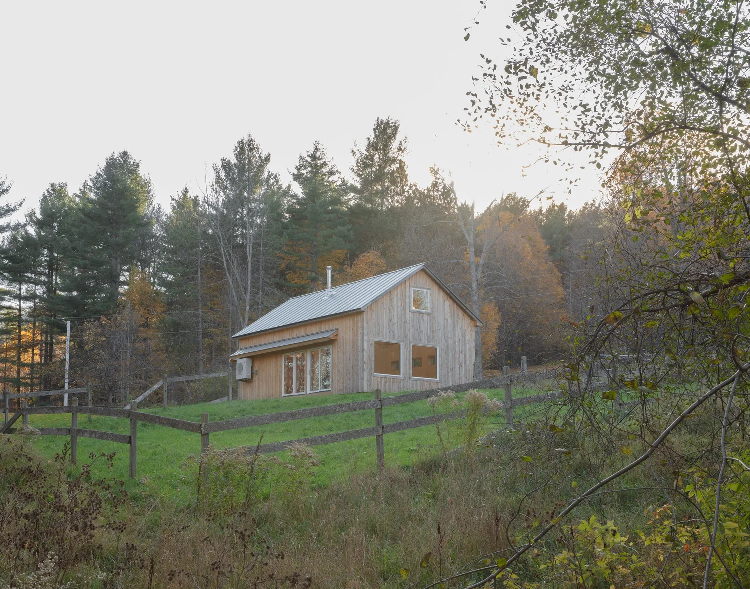 Exterior view of Barnard Barn, a simple gabled structure clad in natural wood siding with a metal roof, set on a grassy slope with a wooden fence and forest backdrop in autumn.