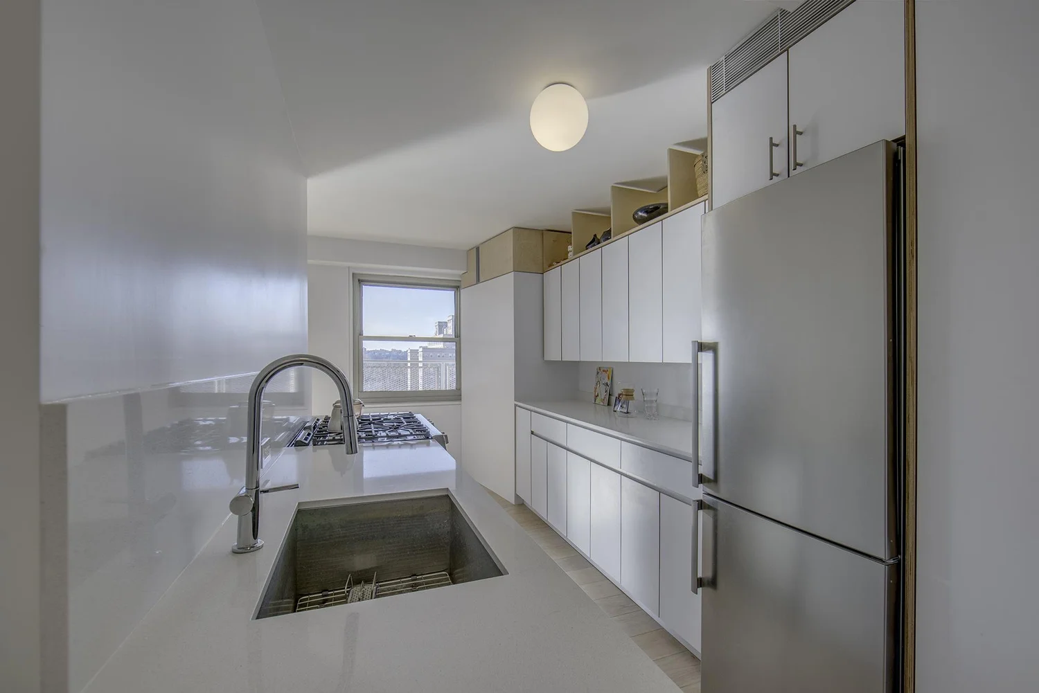 Minimalist kitchen in Harlem Studio featuring white cabinetry, light wood accents, integrated appliances, and a sleek counter with built-in sink and cooktop.