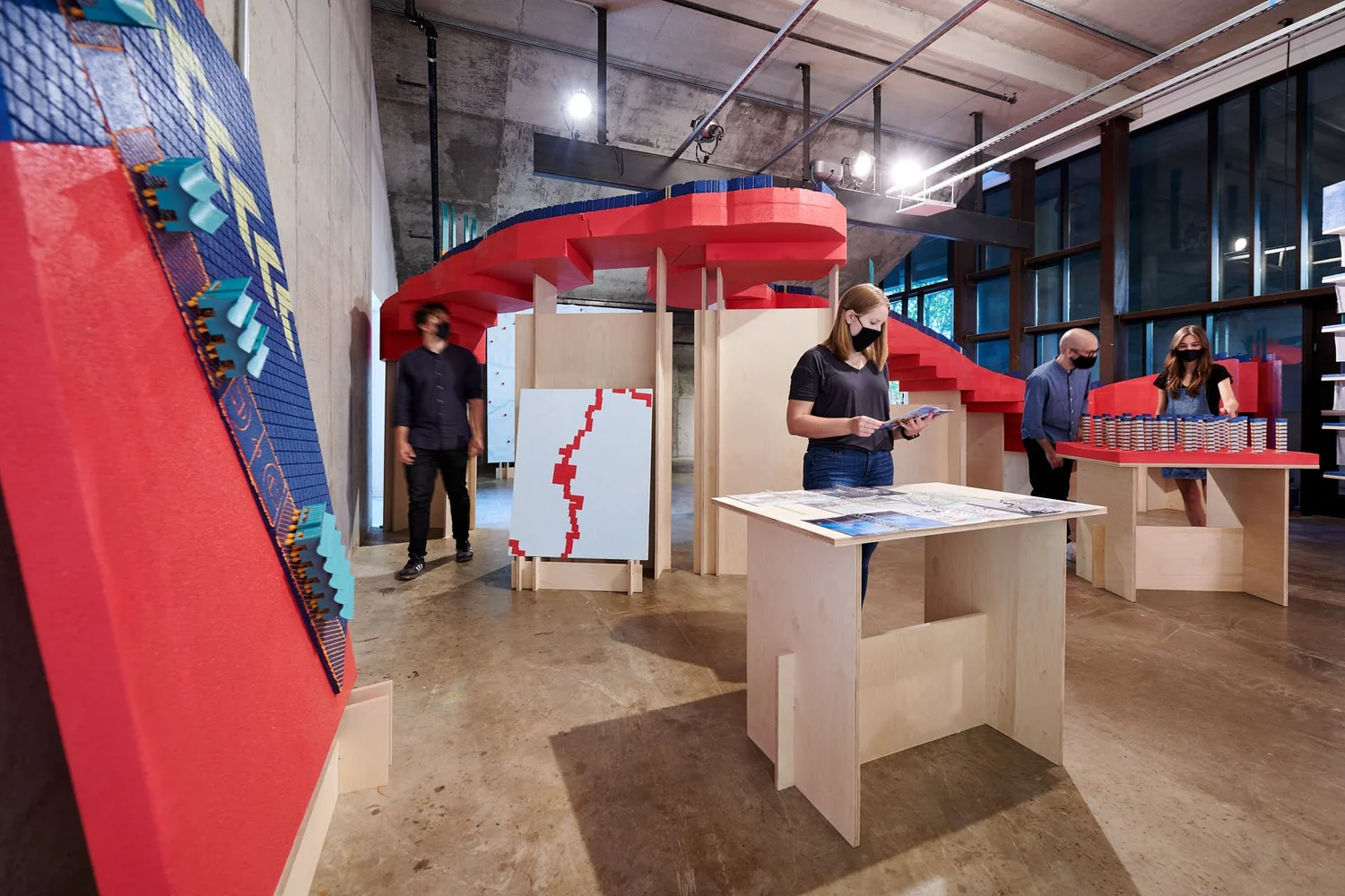 Exhibition view of The Great Lakes Architectural Expedition with red and blue architectural models, wood display tables, and visitors engaging with the installation.
