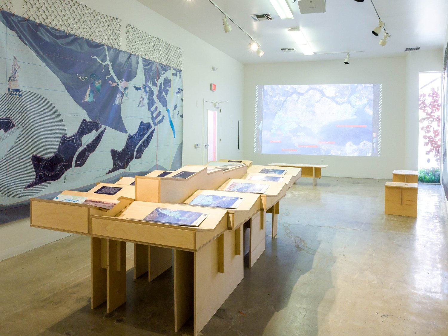 Gallery installation of Territories of Territory Extraction featuring a large wall graphic, stepped wood display with books, and a projected map on the opposite wall.