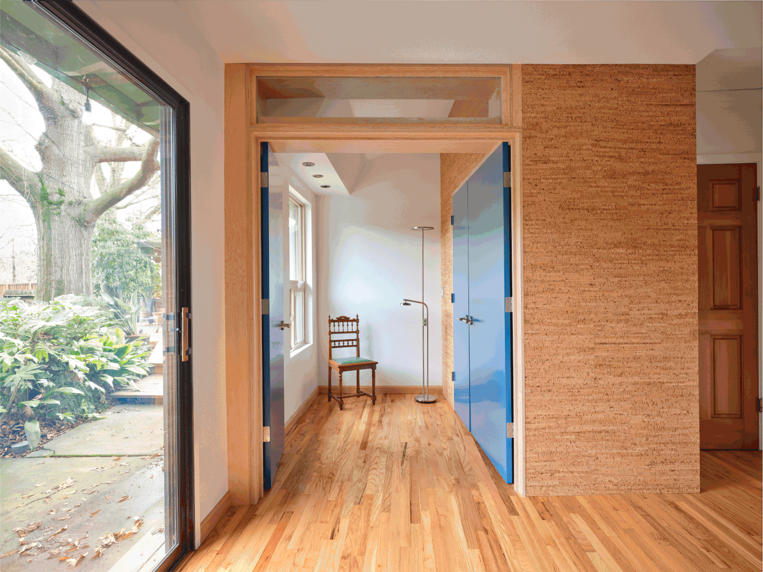 Interior view of Three Cork Walls showing bold blue double doors framed in wood, cork-clad walls, and natural light from a glass sliding door leading to the garden.