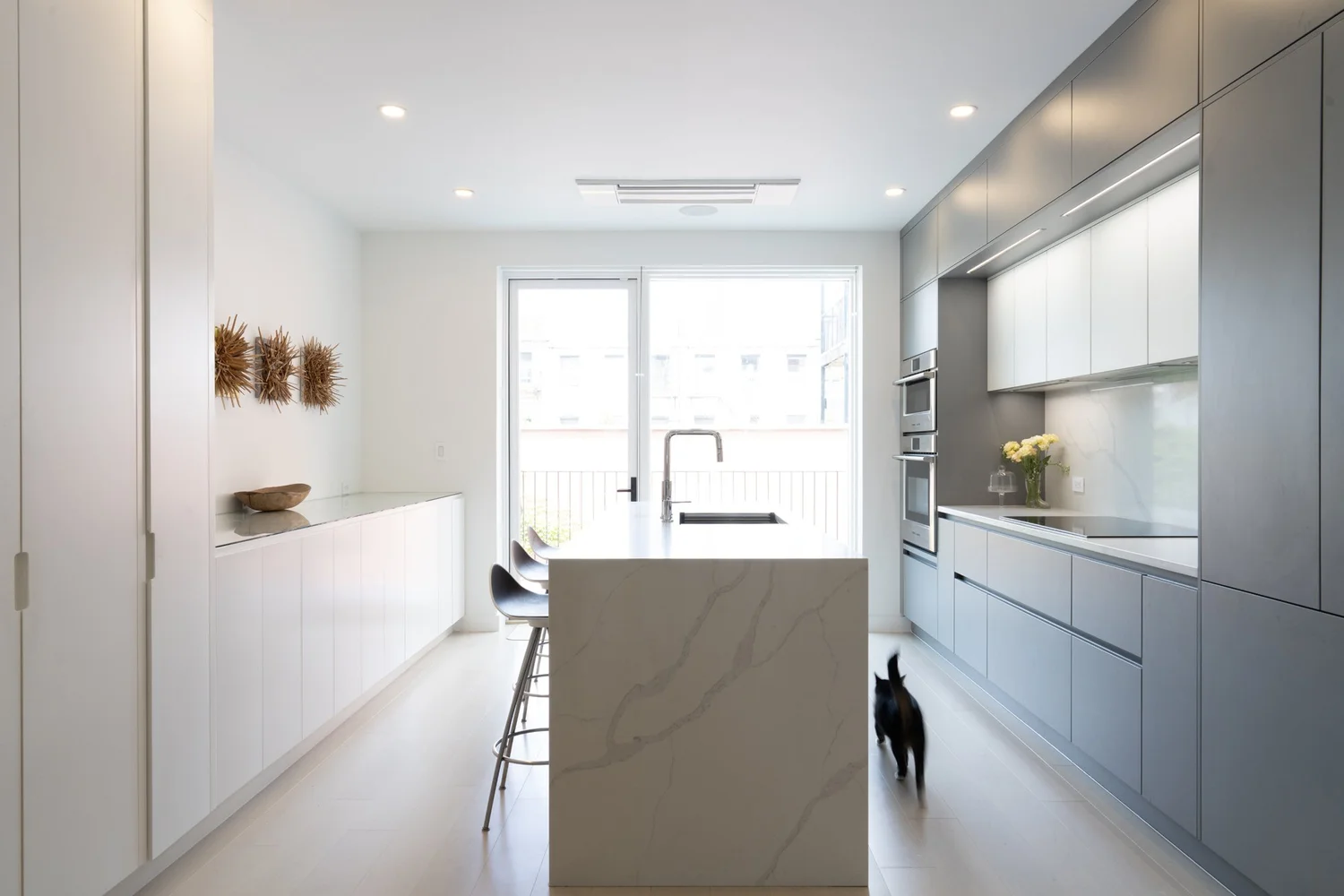 Modern kitchen at Narrow Townhouse with marble island, integrated gray cabinetry, wall ovens, and floor-to-ceiling glass doors opening to a terrace.