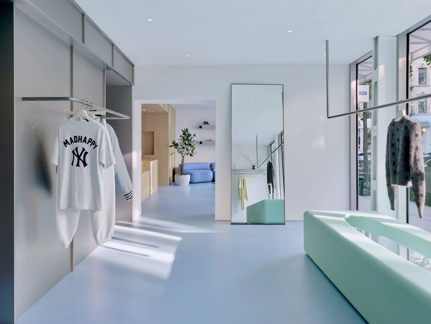 Interior of Madhappy New York Flagship store, showing branded apparel on display, a full-height mirror, sculptural mint-green bench, and lounge area with blue sofa.