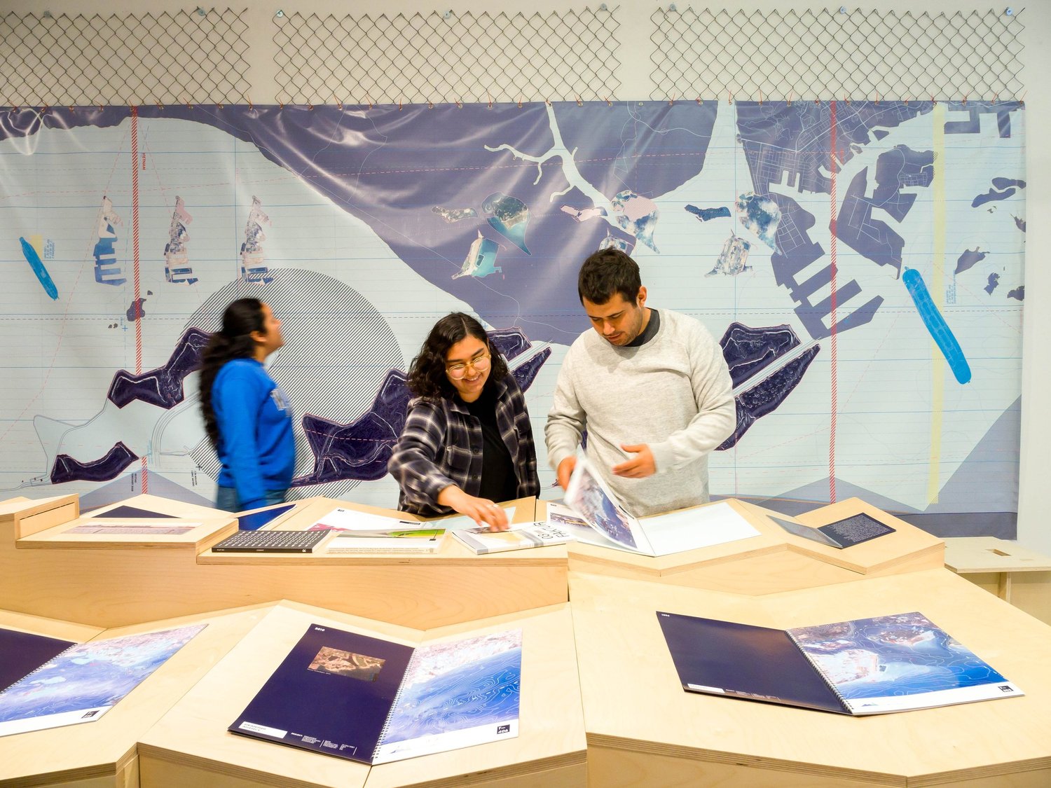 Visitors interacting with research books in the Territories of Territory Extraction exhibition, with a large wall graphic in the background.