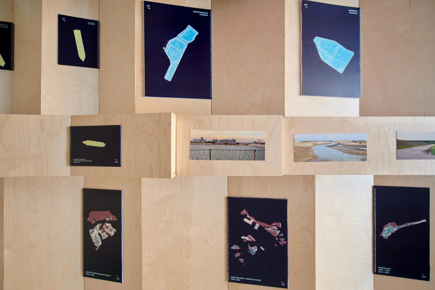 Close-up of Territories of Territory Extraction exhibition showing spiral-bound books with maps and research images displayed on a stepped wood platform.