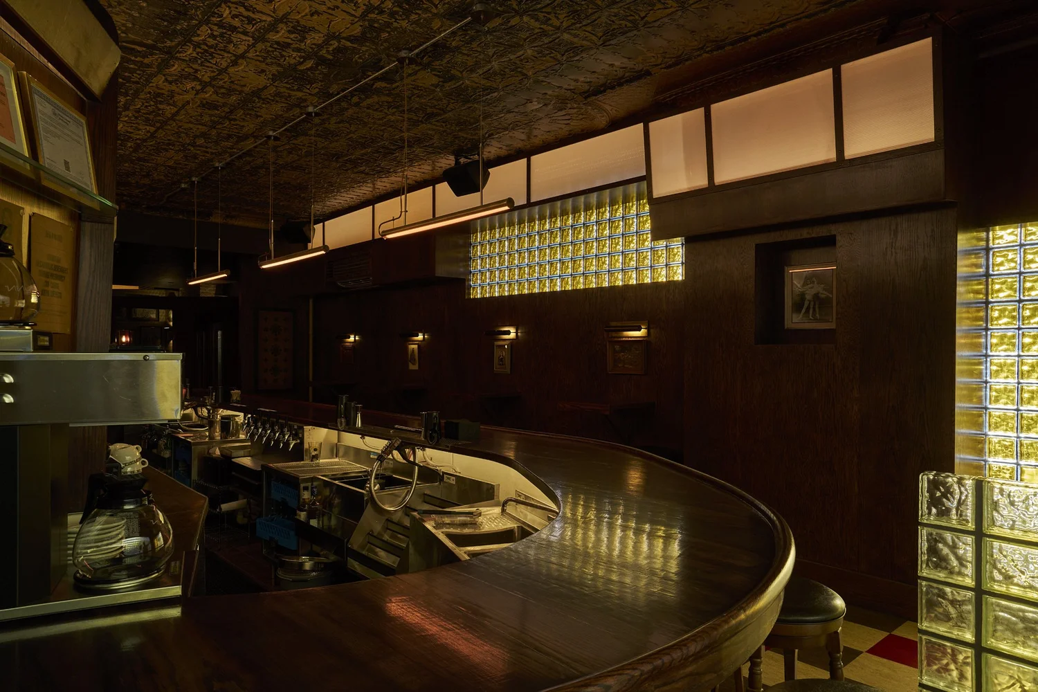 Interior of Mr. Nancy’s bar featuring a curved dark wood counter, vintage glass block walls, tin ceiling, and warm ambient lighting.