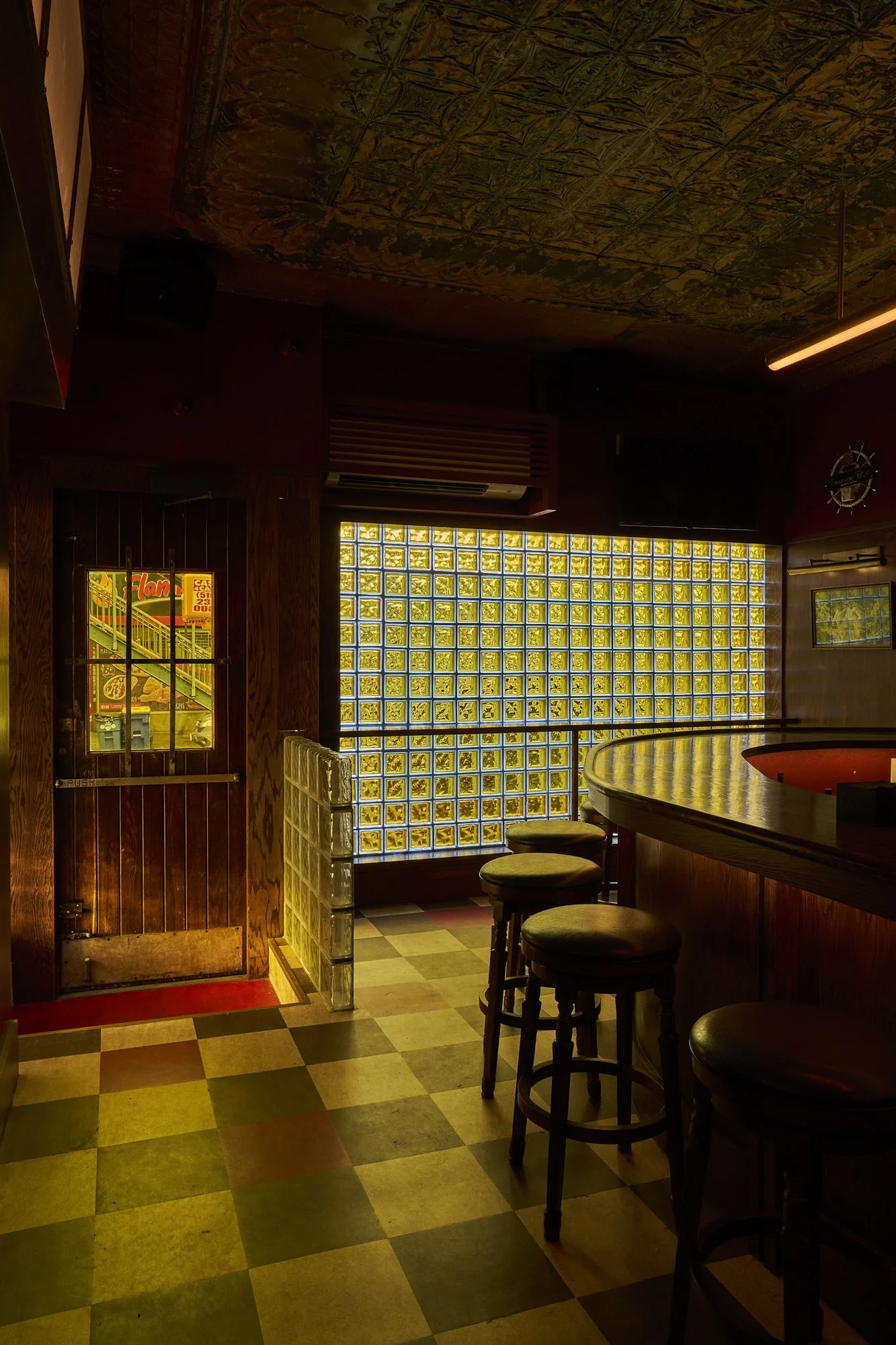 Entry door at Mr. Nancy’s with wooden paneling, checkerboard tile floor, and a backlit glass block wall beside the bar.