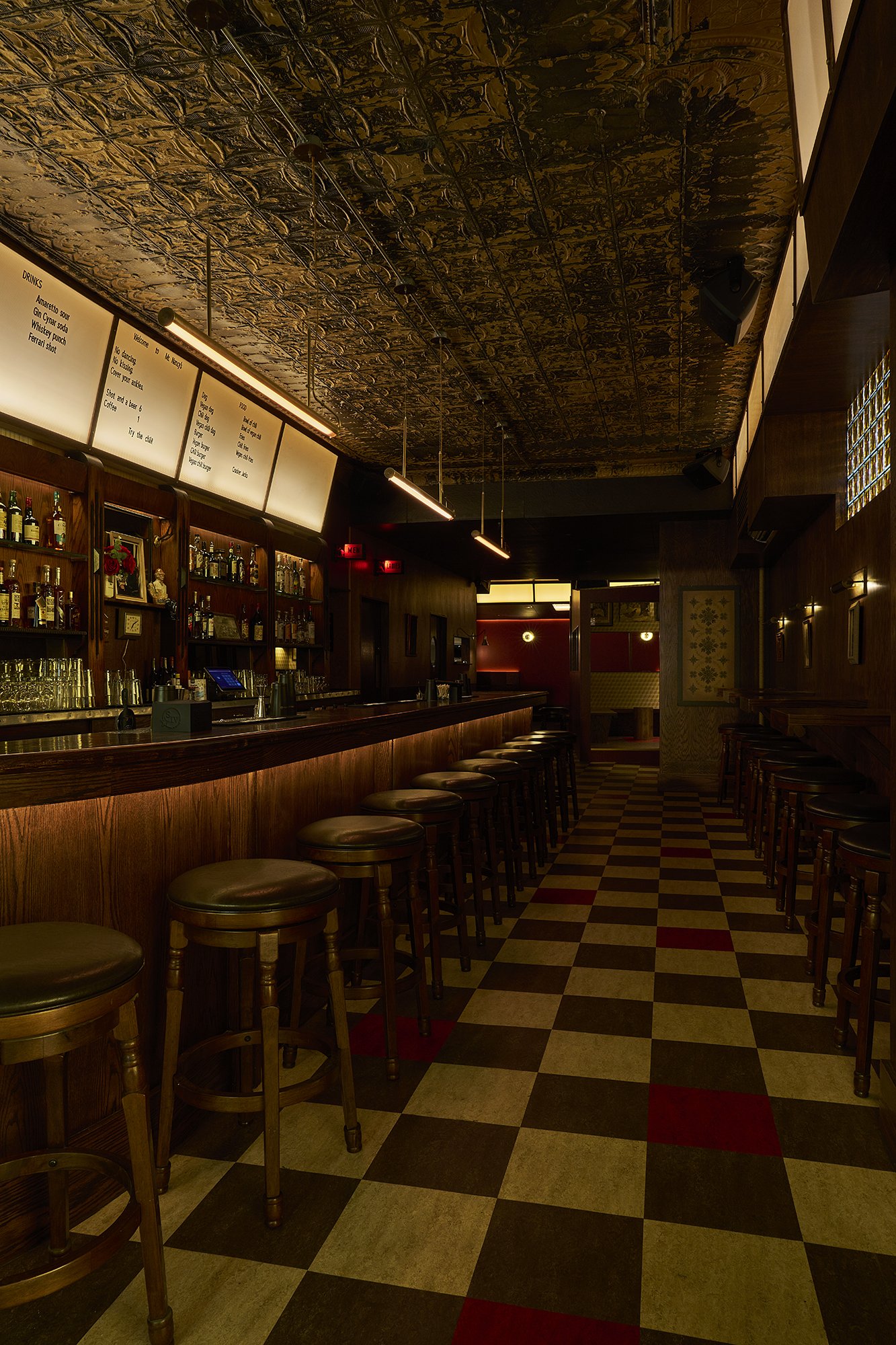 Long perspective view of Mr. Nancy’s bar interior with tin ceiling, illuminated menu boards, checkerboard tile floor, and rows of wooden bar stools.