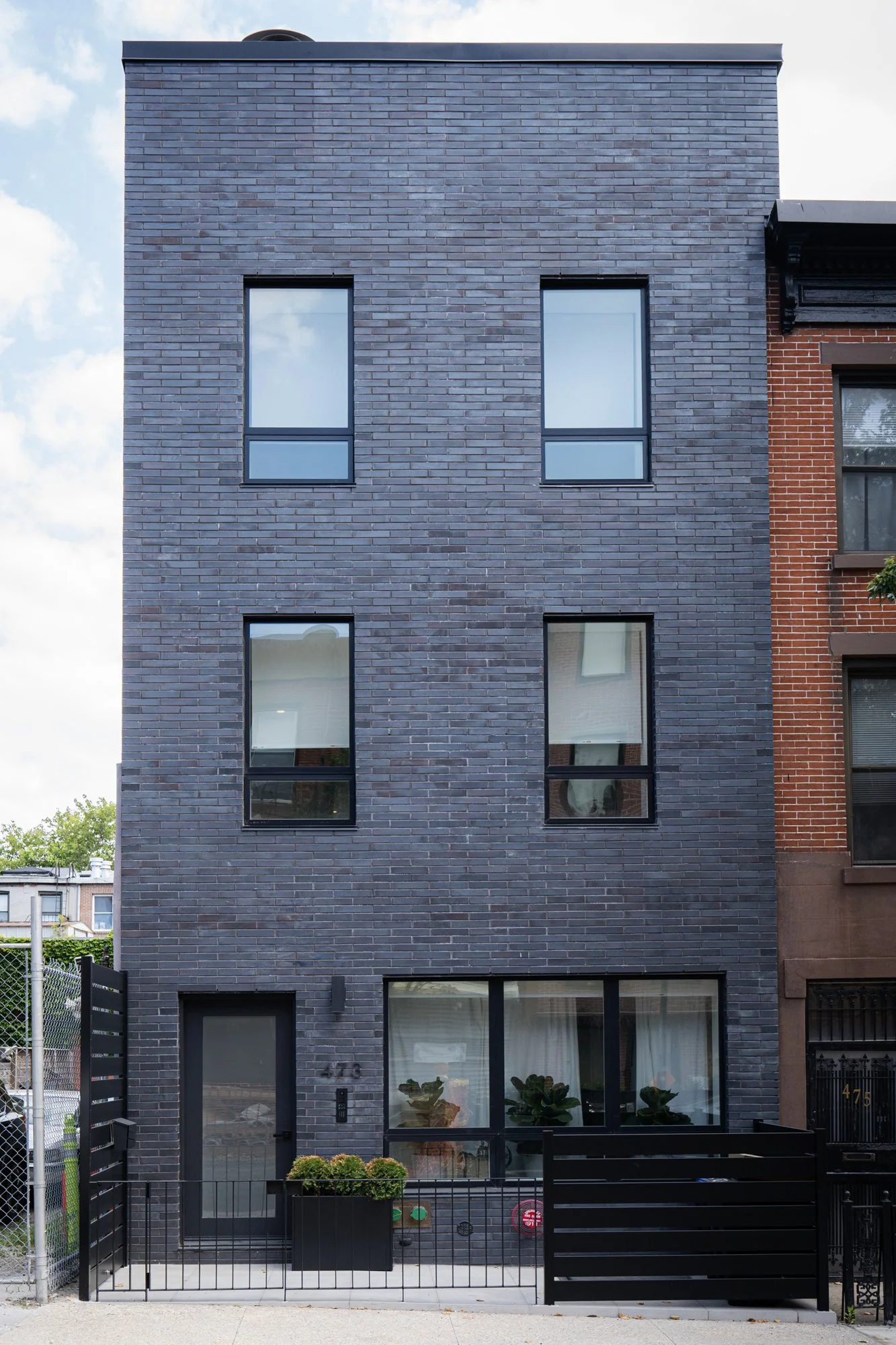Modern three-story townhouse with a dark gray brick façade, large rectangular windows, and a black metal gate in Brooklyn.