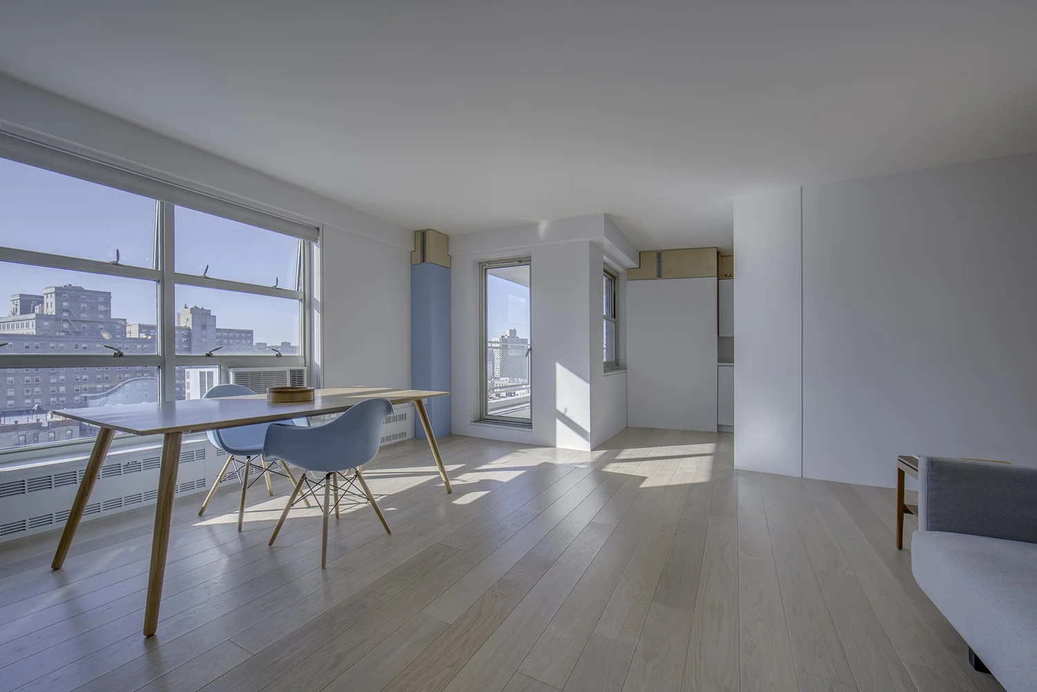 Interior of Harlem Studio featuring a bright, sunlit room with large windows, light wood floors, a simple wood desk, and a blue Eames chair. Minimal furniture emphasizes openness and natural light.