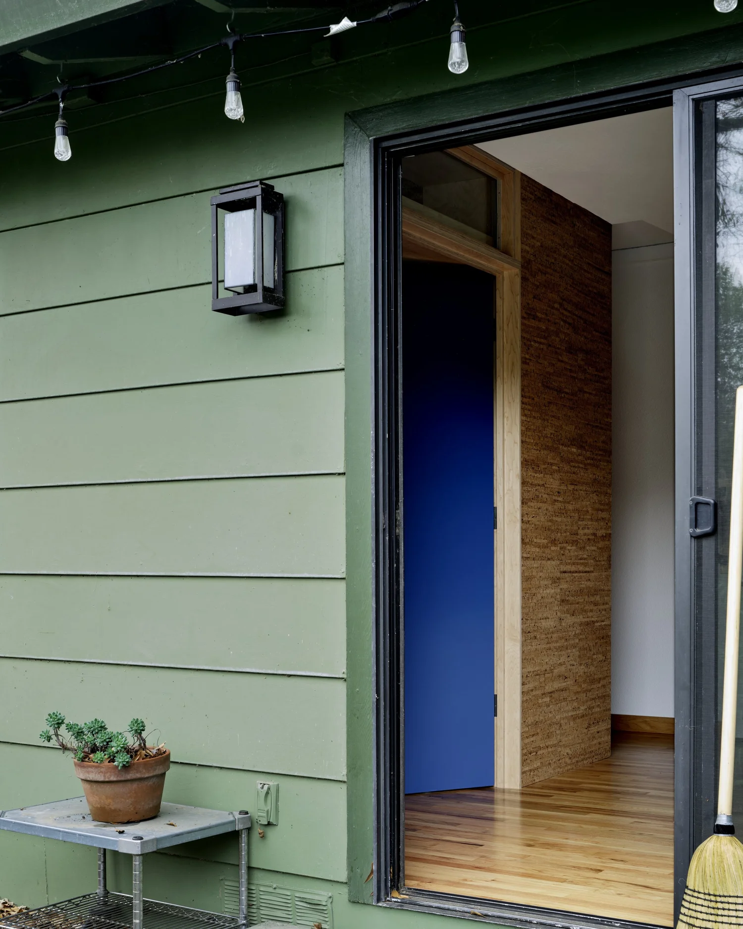 Exterior entry of Three Cork Walls with green siding and a glimpse inside to blue doors framed by cork walls.