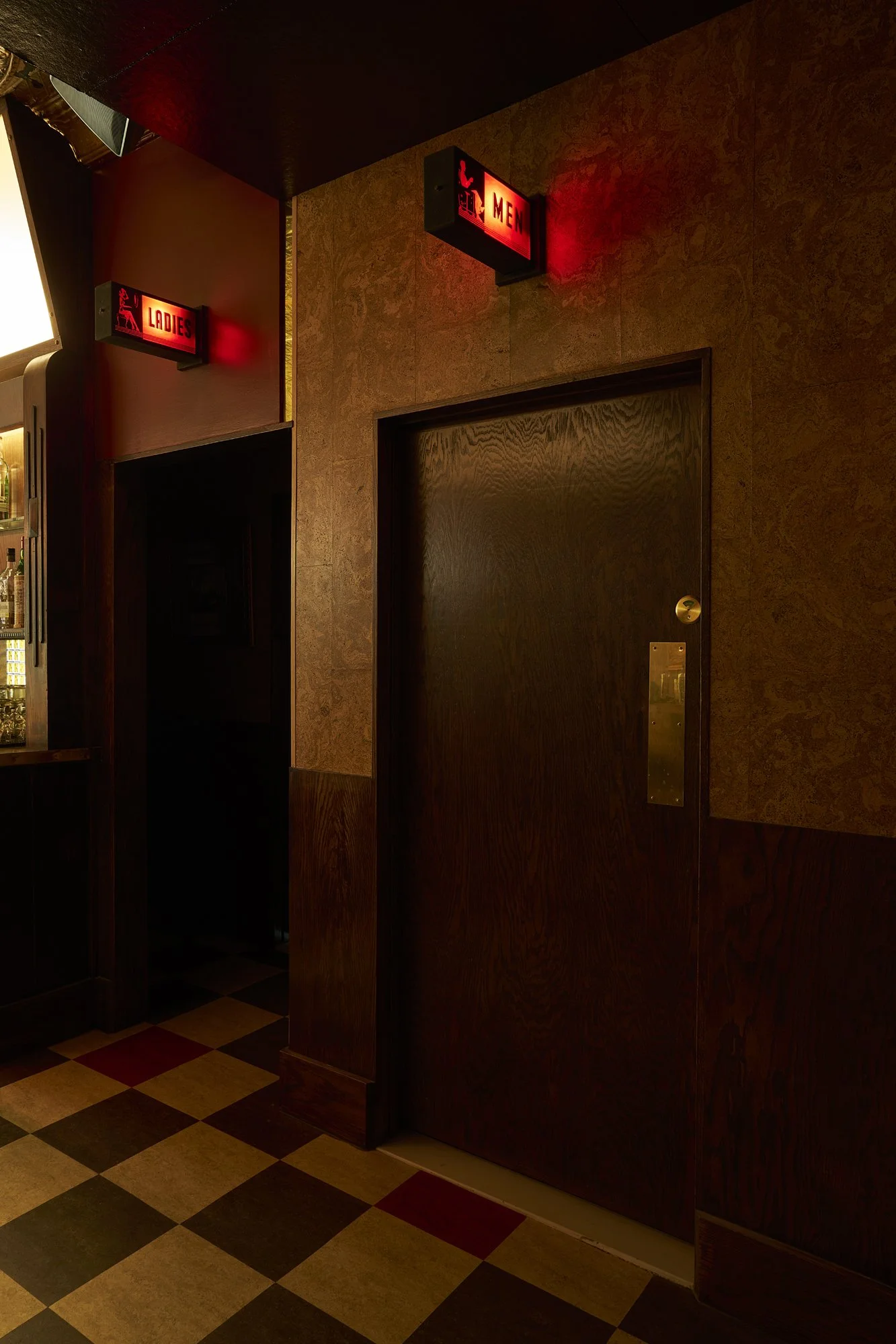 Bathroom door at Mr. Nancy’s with dark wood paneling, illuminated red exit sign, and checkerboard tile floor.