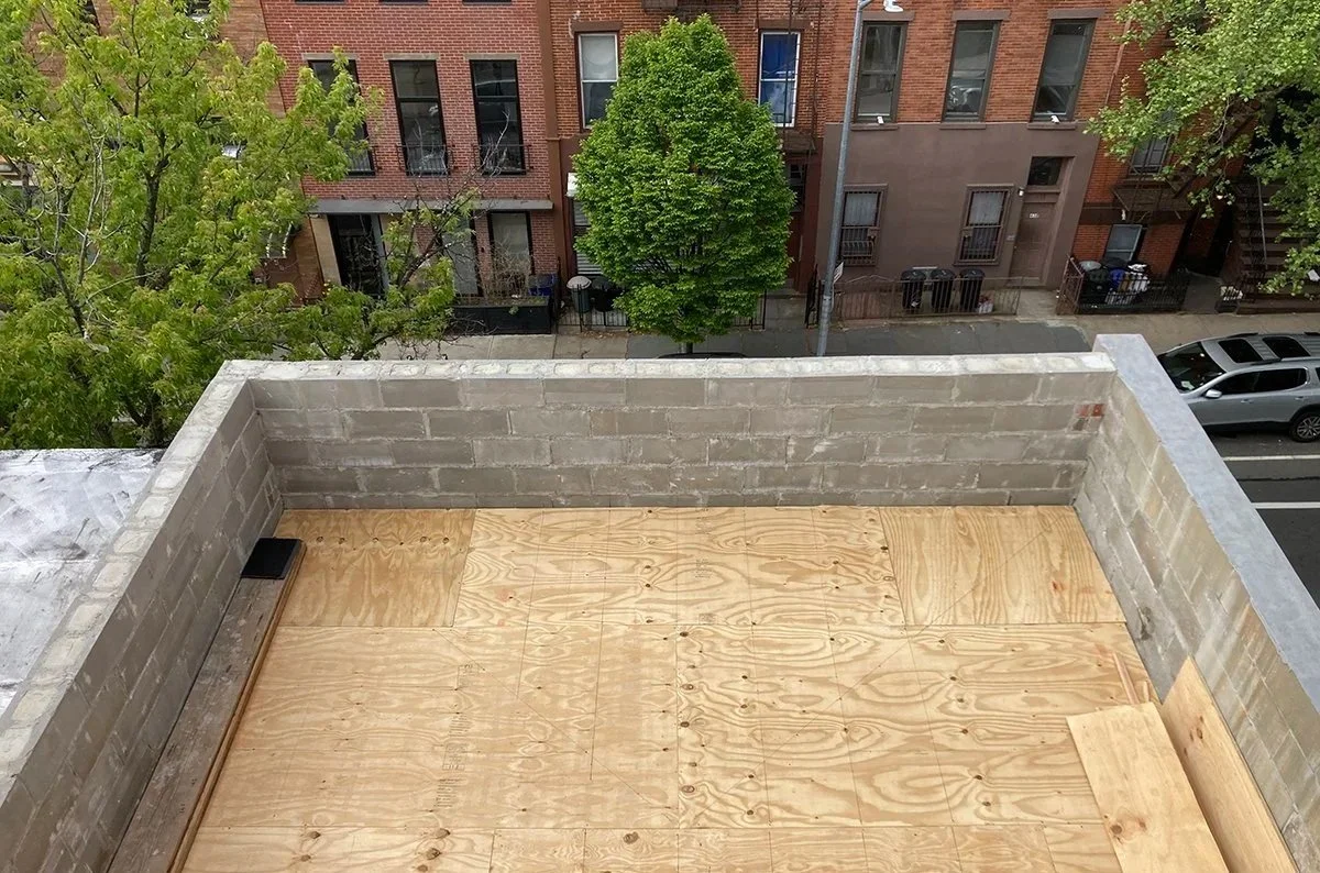 Rooftop view showing a newly built plywood deck with low brick walls overlooking a tree-lined street.