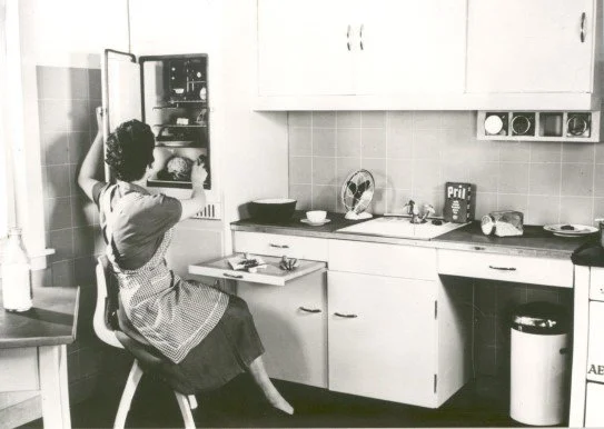Black-and-white photograph of the Frankfurt Kitchen designed by Margarete Schütte-Lihotzky, showing a woman seated on a stool working within a compact, efficient kitchen with built-in cabinetry and integrated appliances.