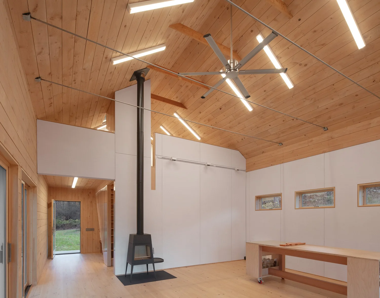 Interior view of Barnard Barn showing wood-paneled ceiling and walls, a tall black wood stove with chimney pipe, sliding barn-style track door, and clerestory windows bringing in natural light.